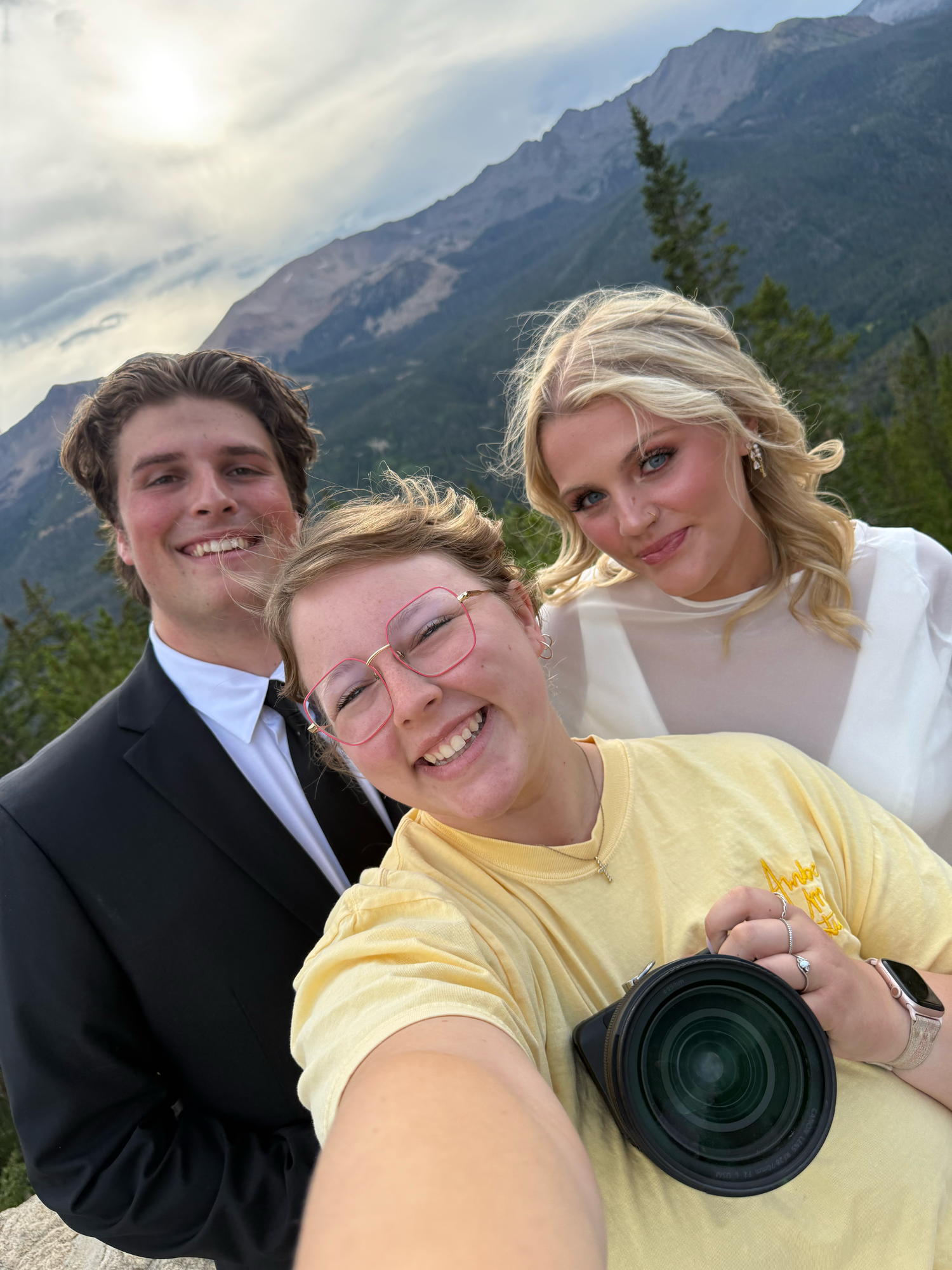 Three smiling people pose for a selfie on a mountain overlook, one holding a camera with a large lens, with rugged peaks behind them.