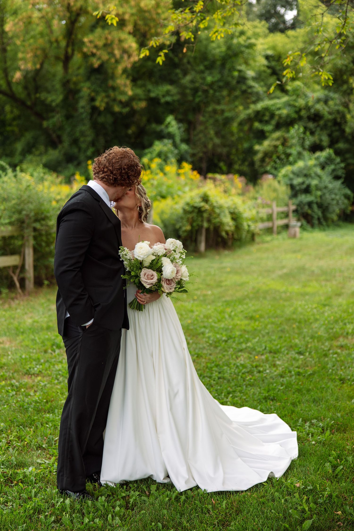 Bride in a flowing white gown holds a pink and white bouquet while embracing her groom in a black suit on a lush green lawn.
