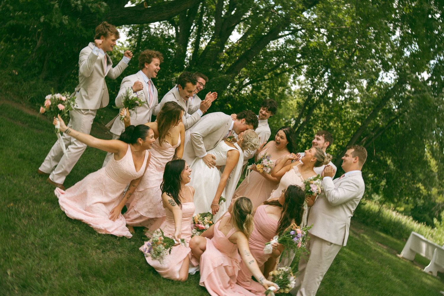 Bridal party in blush dresses and tan suits laughing together on a grassy hillside under shady trees while holding bouquets.