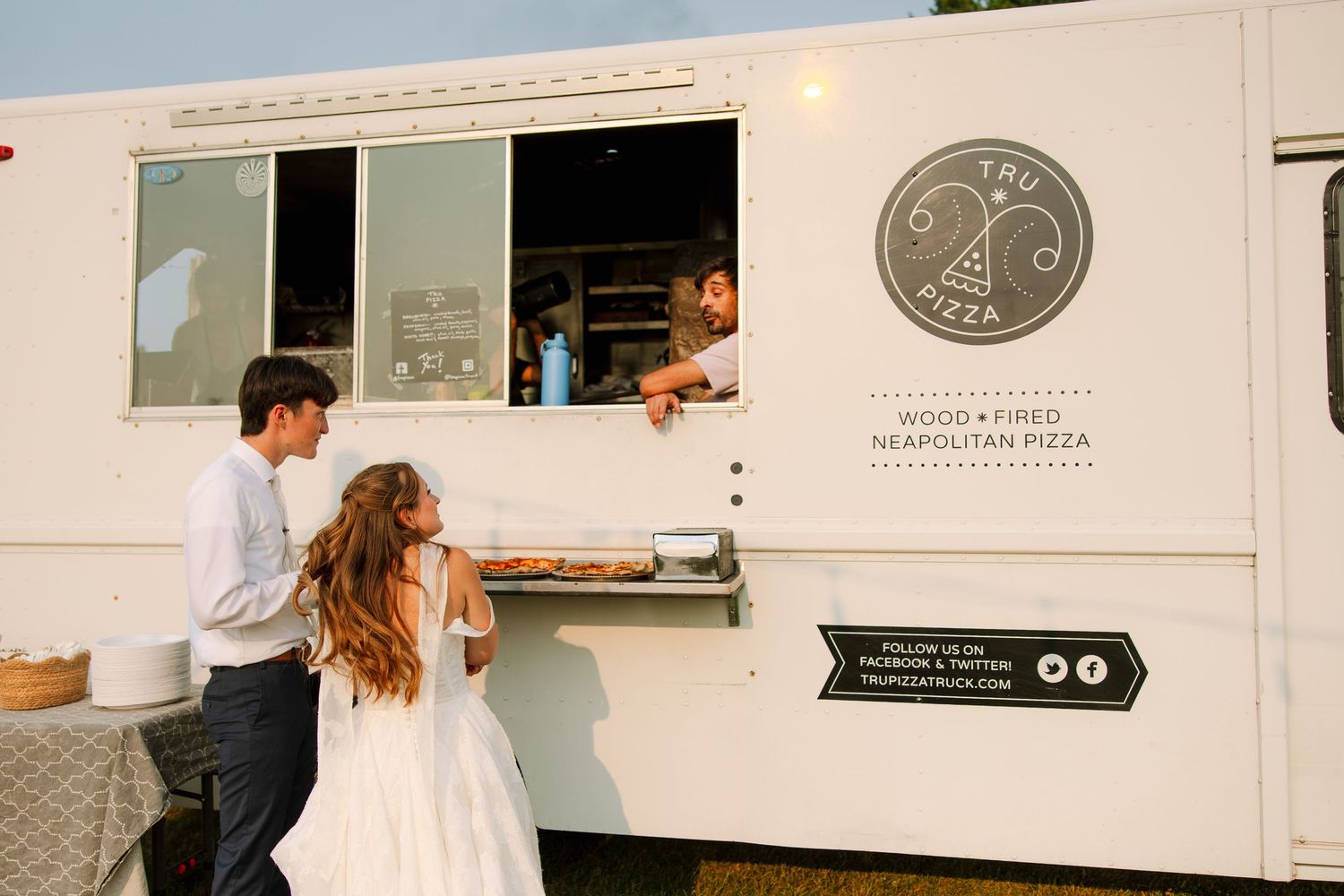 Bride and groom order wood-fired Neapolitan pizza from a white pizza food truck at their outdoor wedding reception.