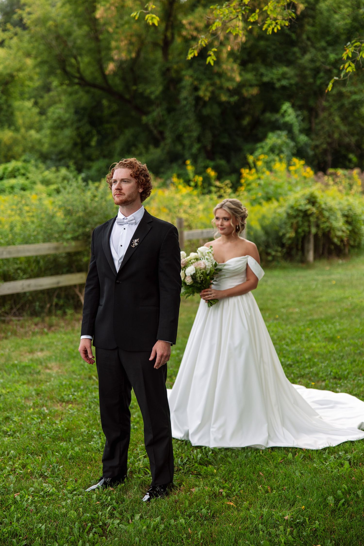 Bride in a white off-shoulder gown holding a bouquet stands behind a groom in a black suit and bow tie during an outdoor garden first look.
