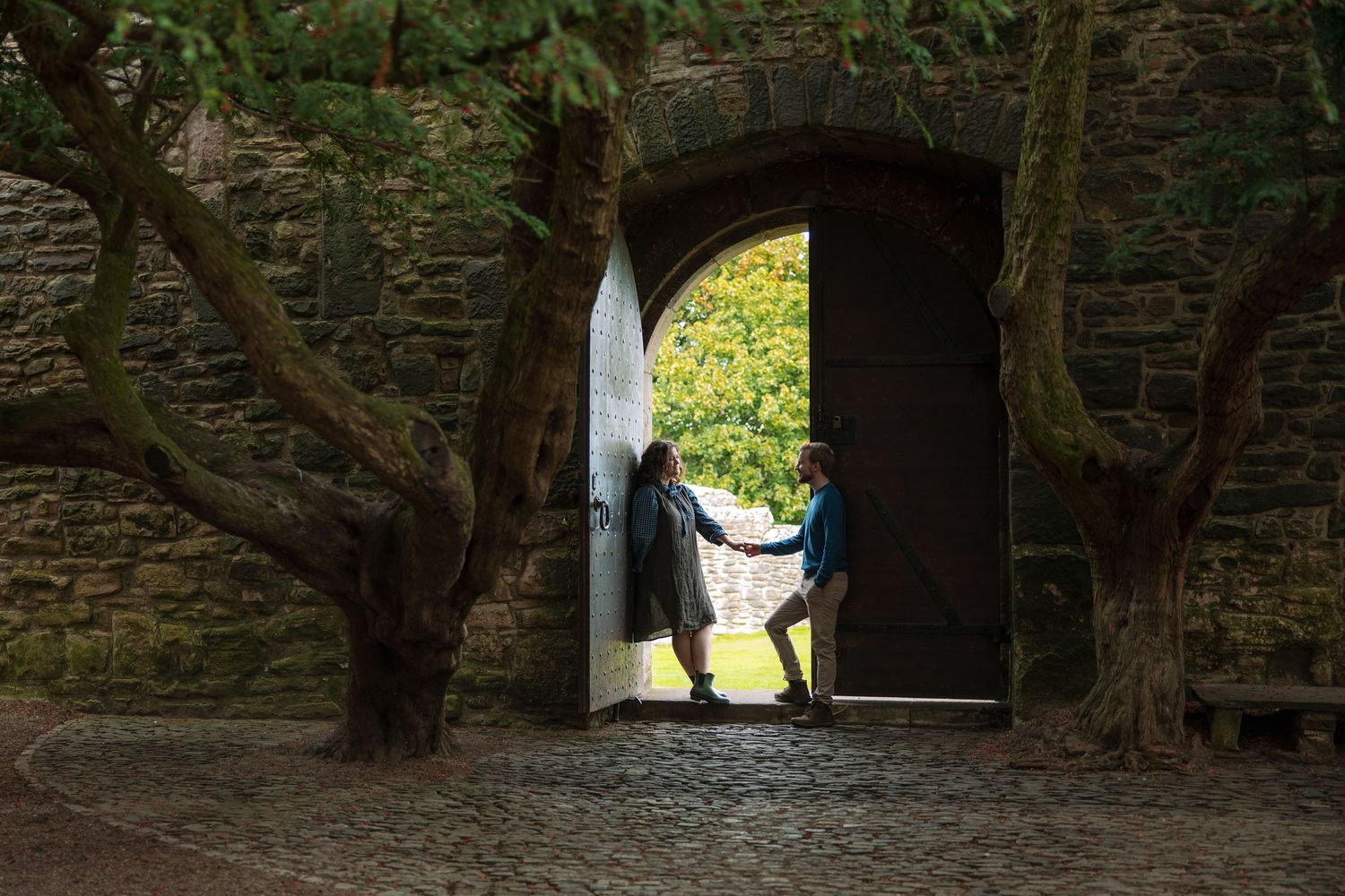 A couple stands holding hands in a stone archway beside gnarled trees, with a sunlit green landscape glowing beyond.