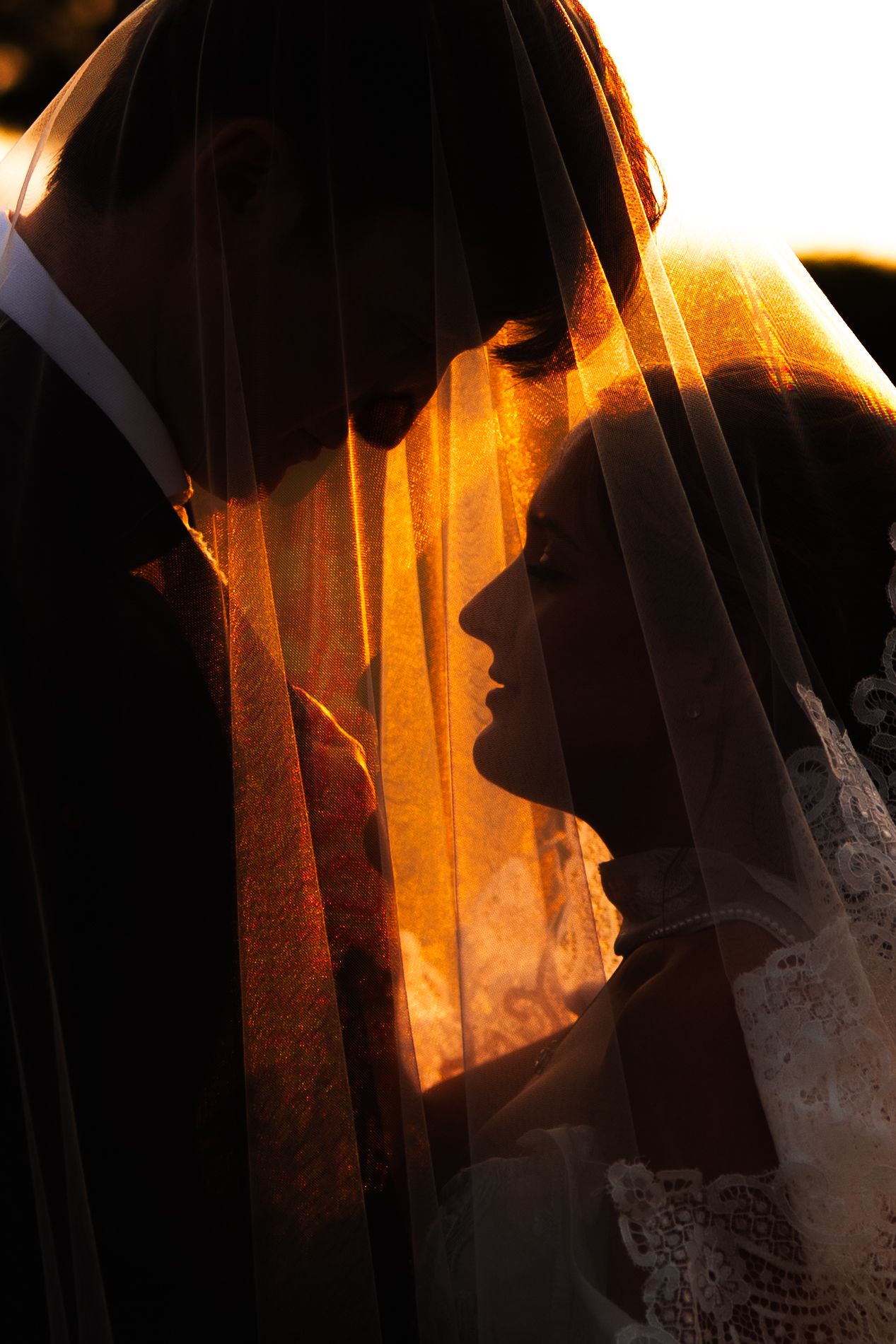 Silhouetted bride and groom stand forehead to forehead beneath a glowing veil, lit by warm orange sunset light.