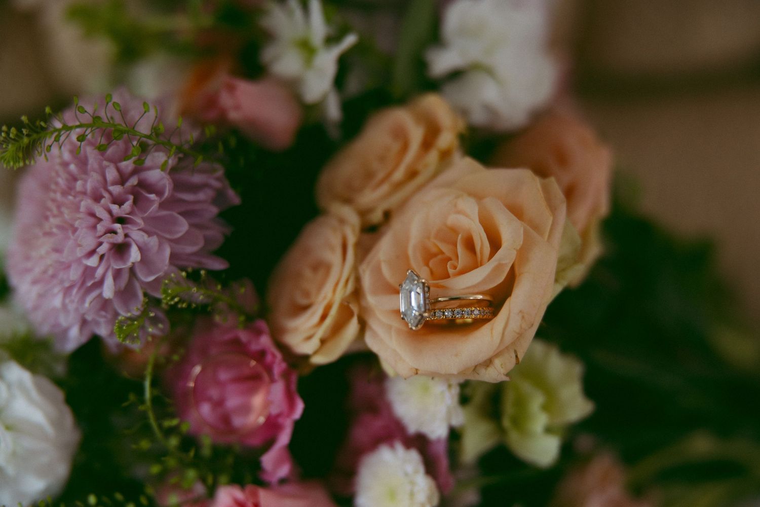 A diamond engagement ring and gold wedding band rest inside a peach rose in a bridal bouquet of pink and white flowers.