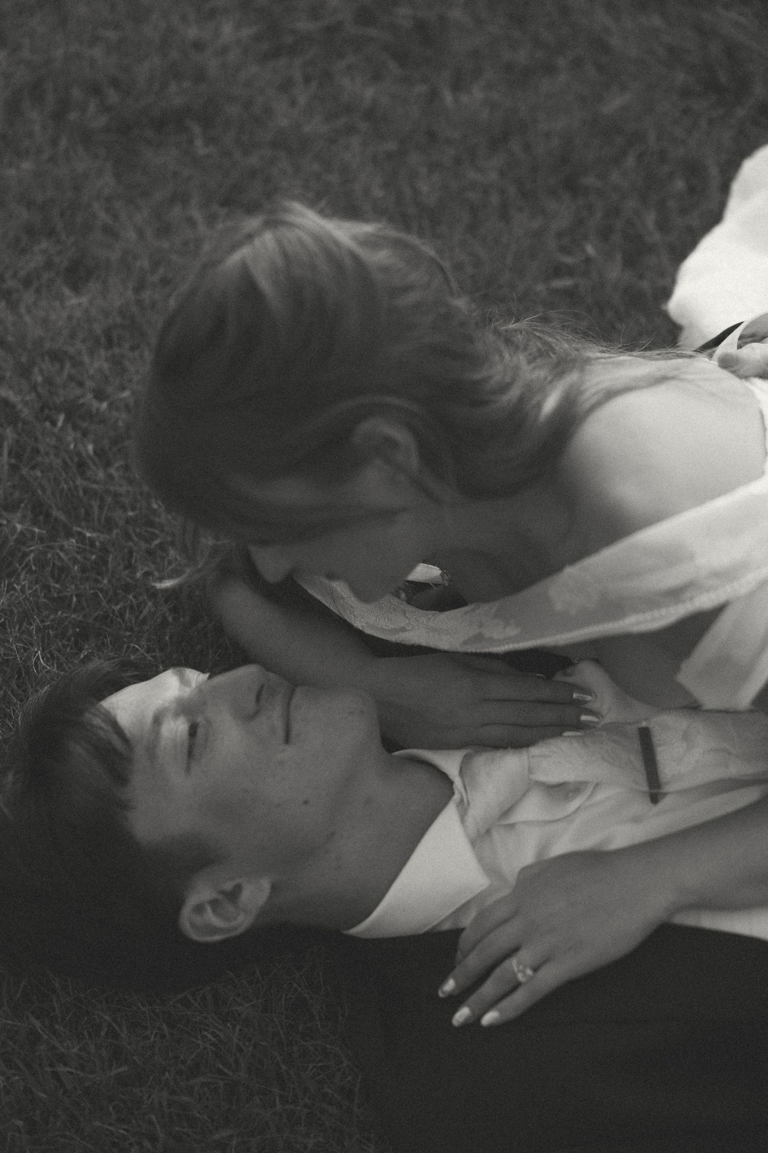 Black and white photo of a bride leaning over a groom as he lies on the grass, wedding rings visible on their hands.