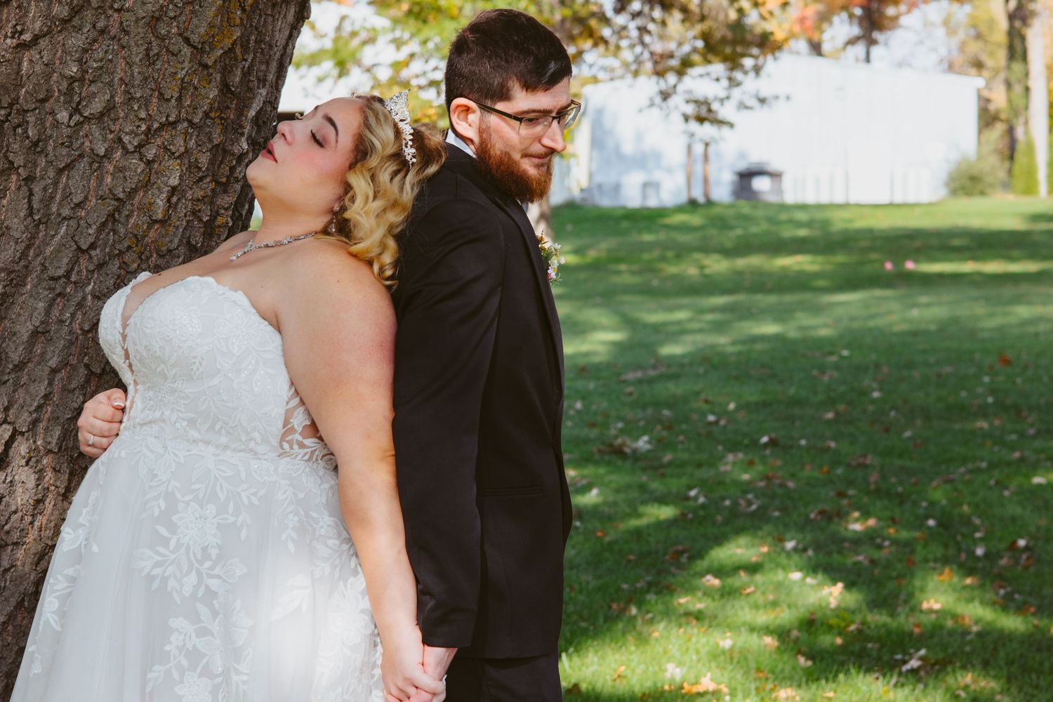 Bride in a lace gown leans her head against a tree trunk while the groom in a dark suit and glasses stands beside her.