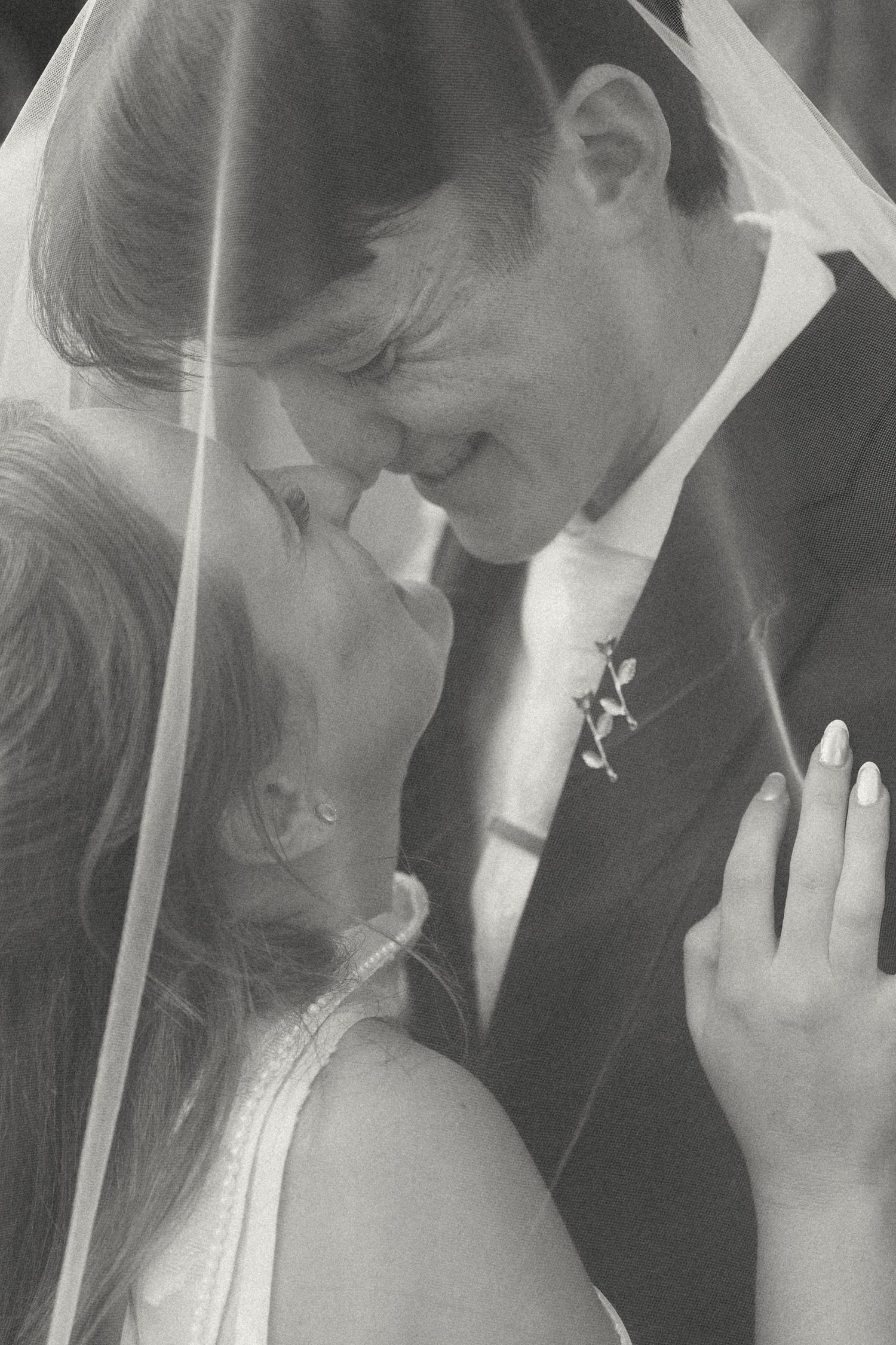 Black and white photo of a bride and groom nose to nose under a sheer veil, smiling during an intimate wedding moment.
