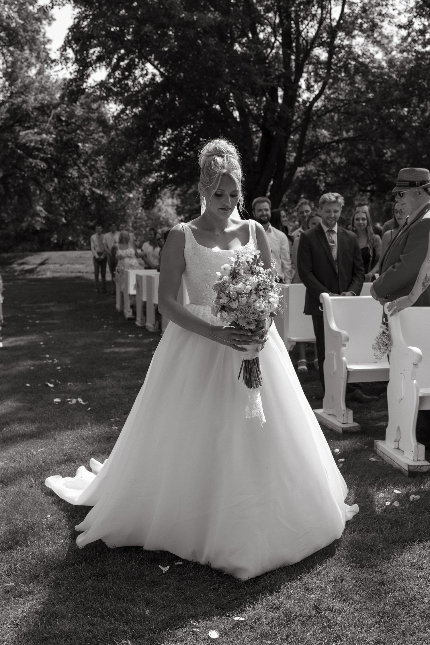 Black and white photo of a bride in a flowing gown holding a bouquet while walking an outdoor garden ceremony aisle past seated guests.