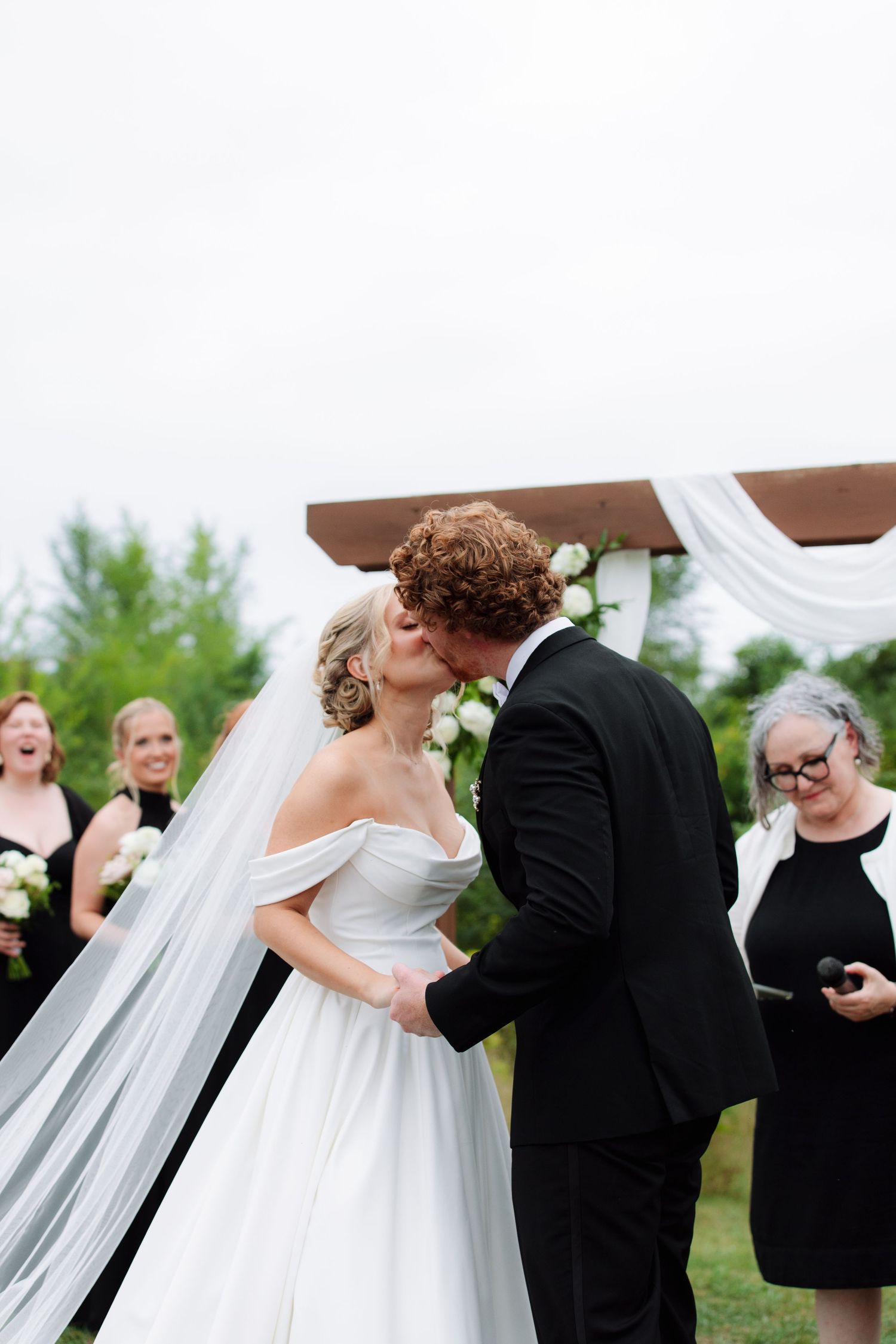 Bride and groom share their first kiss under a white rose-draped arch as bridesmaids and the officiant look on.