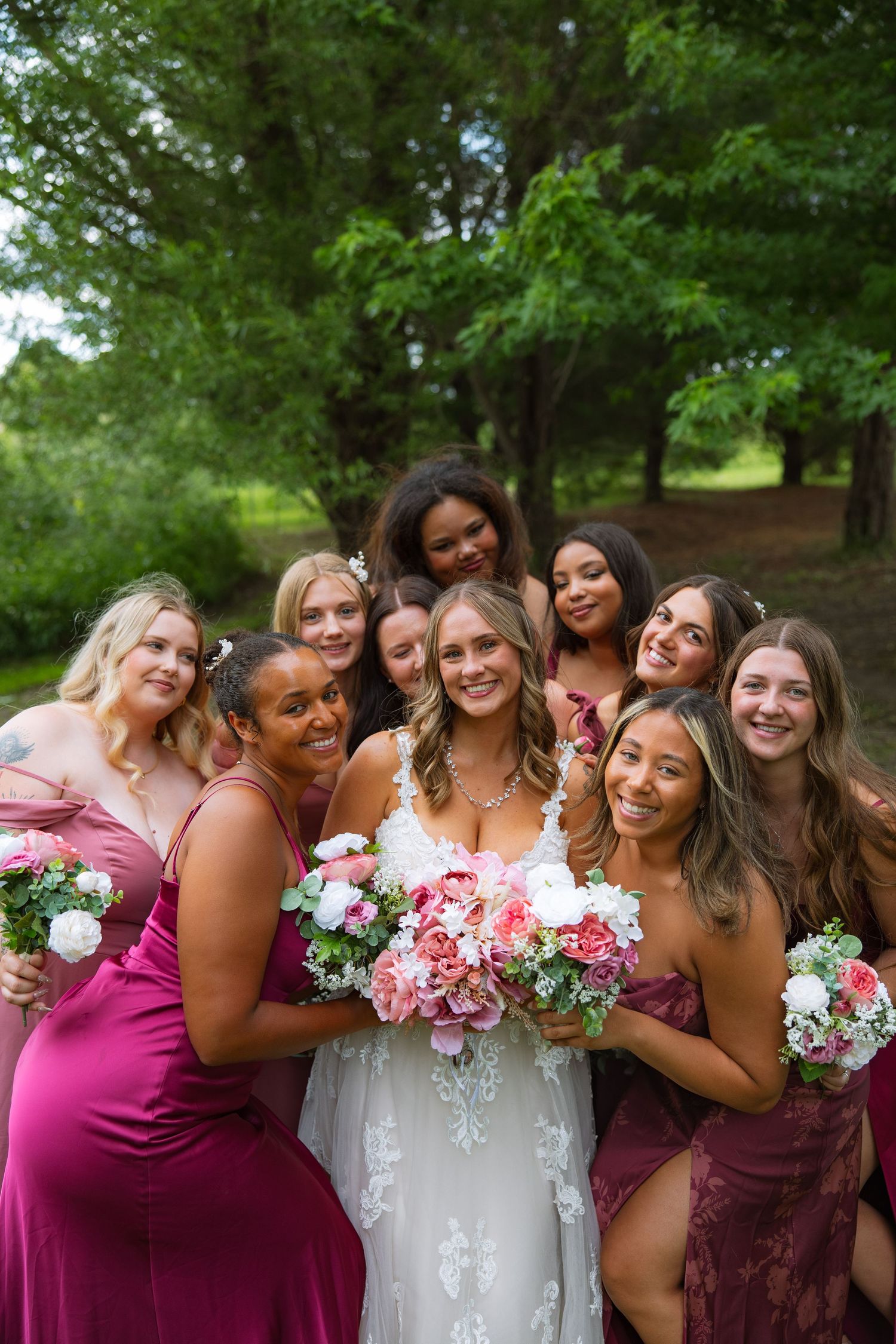 Smiling bridesmaids in magenta and burgundy dresses gather closely around the bride, holding pink and white bouquets outdoors.