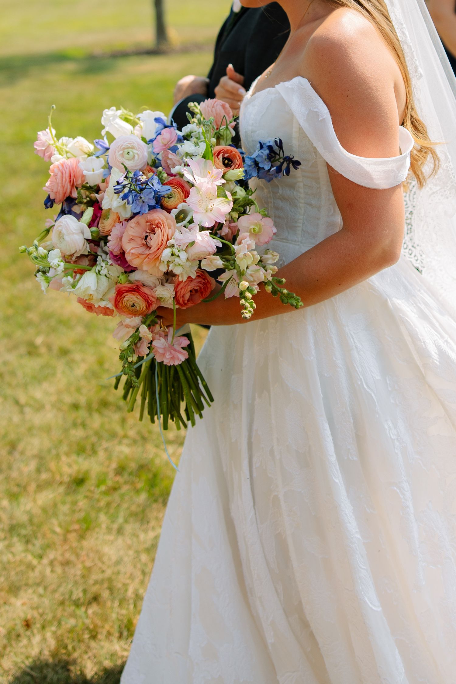 Bride in an off-shoulder white gown holds a colorful wildflower bouquet of peach, pink, and blue blooms on a sunlit lawn.