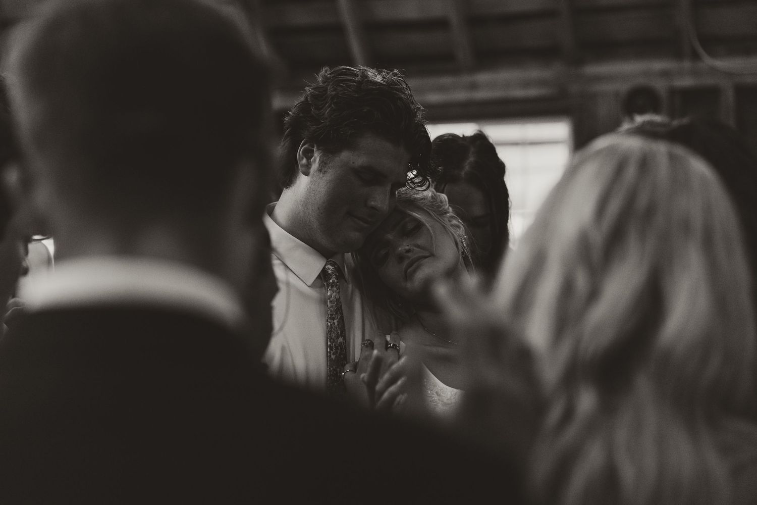 Black and white photo of a groom resting his cheek against the bride's head during a quiet moment at the wedding reception.