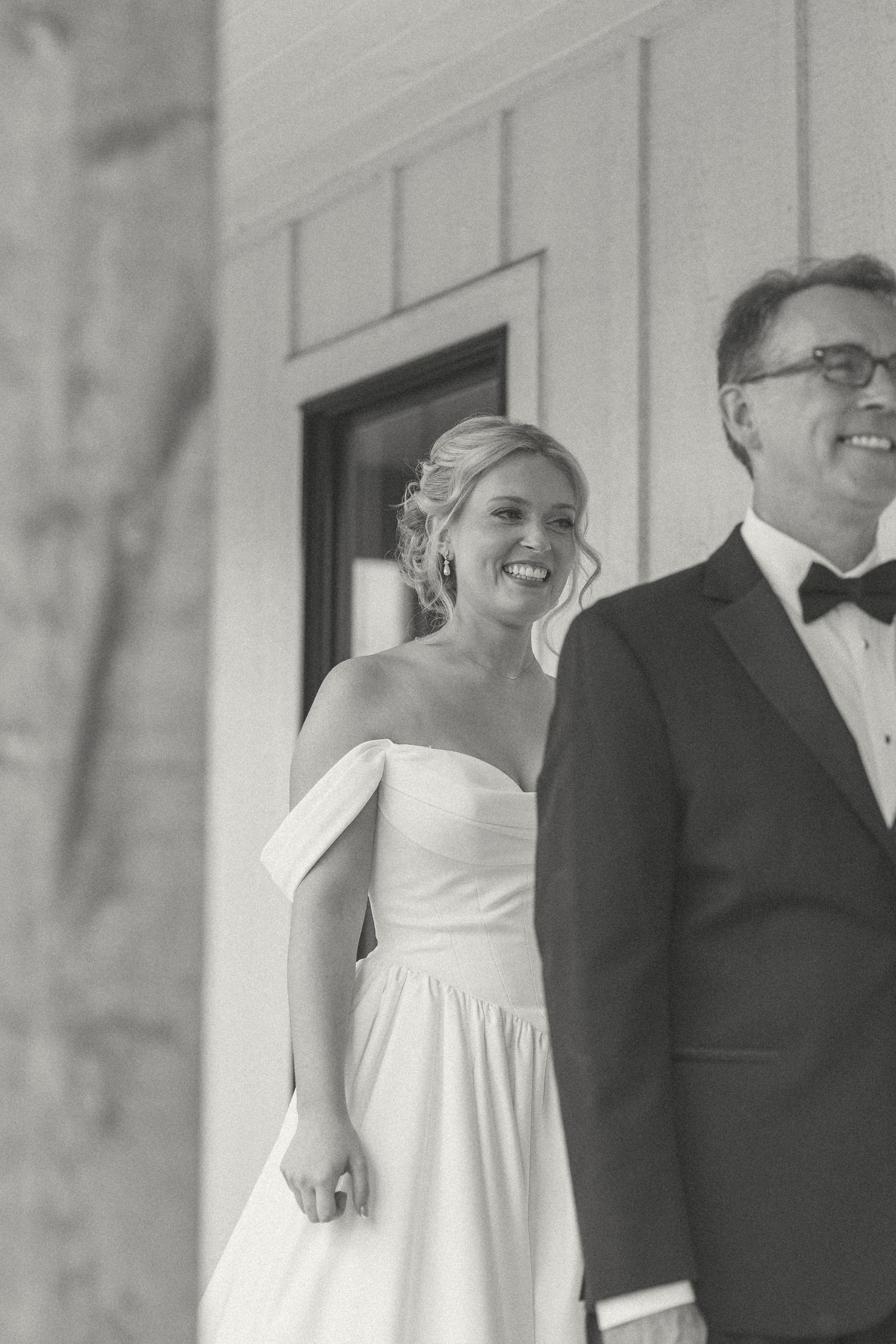 Smiling bride in an off-the-shoulder white wedding gown stands beside a groom in a tuxedo and bow tie in a black and white photo.