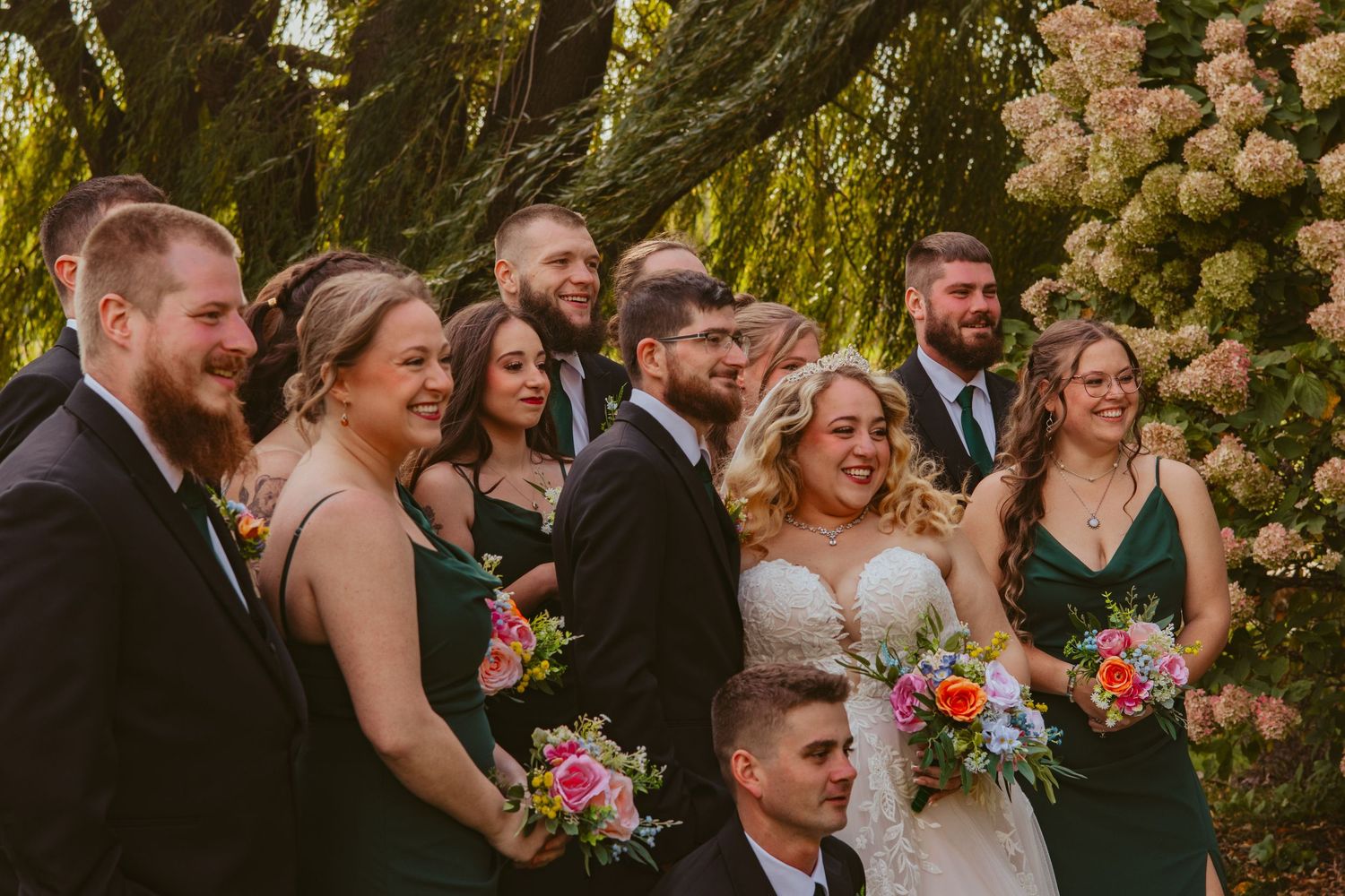 Smiling bride and groom pose with bridesmaids in green dresses and groomsmen in dark suits holding colorful bouquets outdoors.