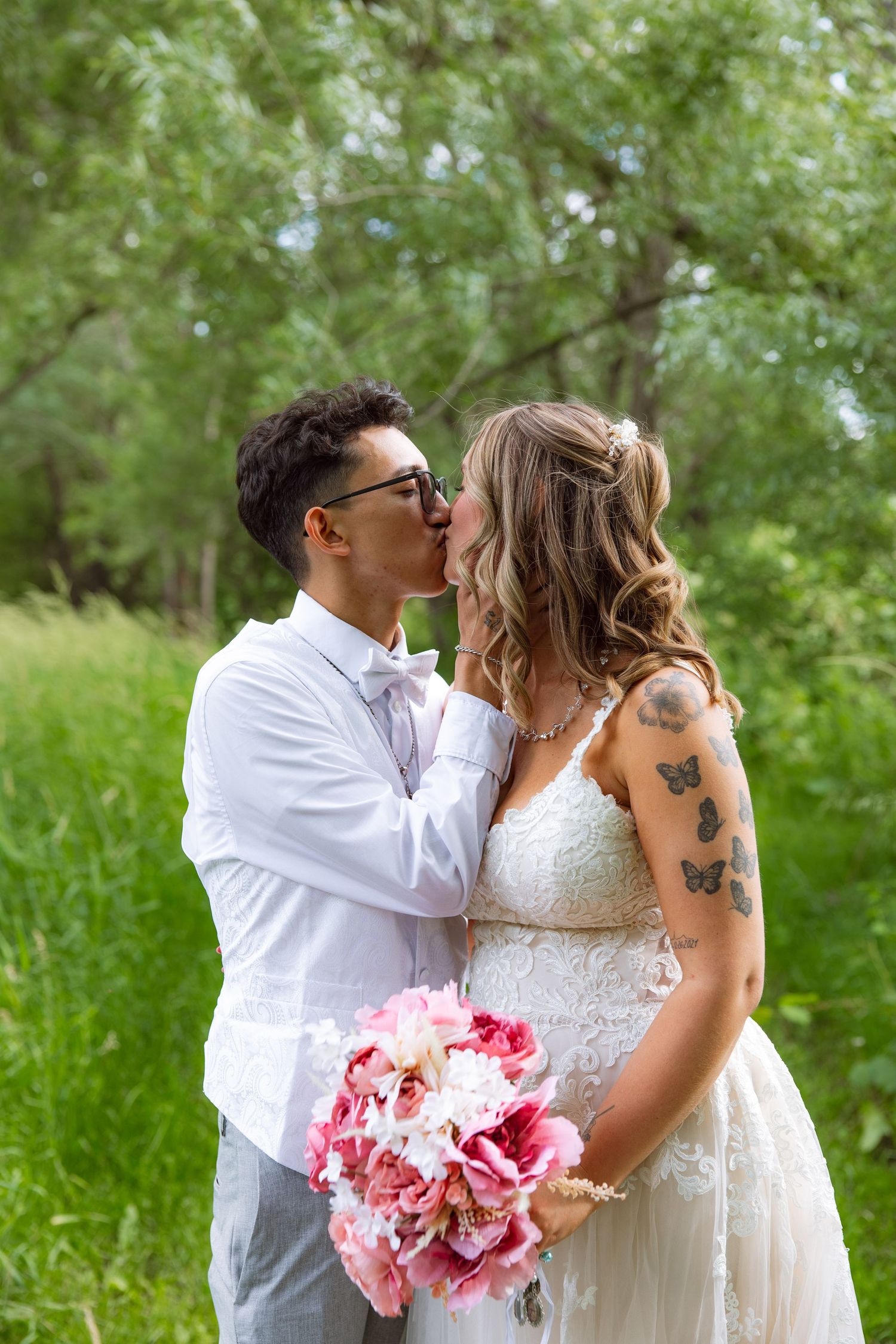 A groom in glasses and a bow tie kisses his bride in a lace wedding gown holding a pink bouquet in a green meadow.