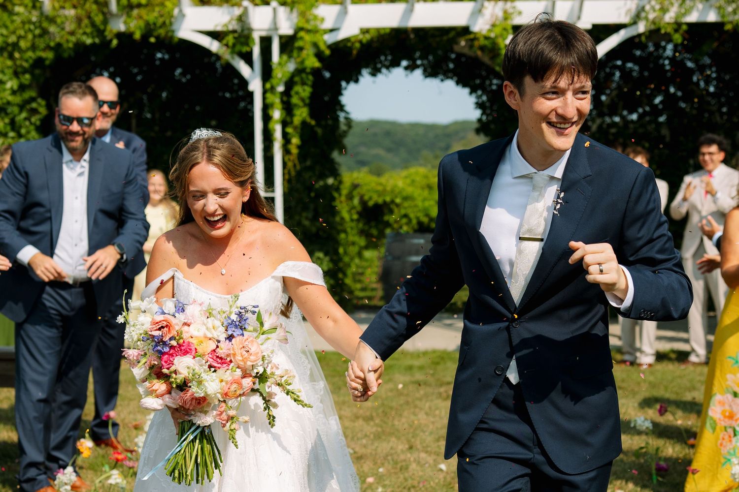 A smiling bride holding a colorful bouquet and a laughing groom hold hands as they walk back down the aisle after their ceremony.