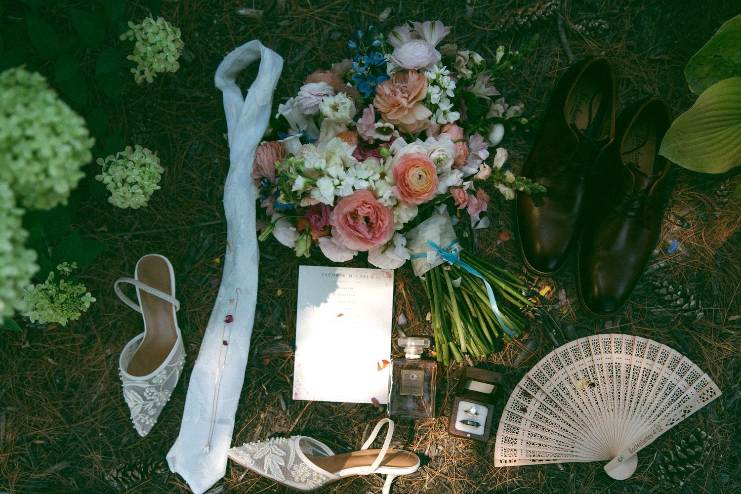 Flat lay of a bridal bouquet, lace heels, groom's shoes, folded fan, veil and invitation card on a mossy forest floor.