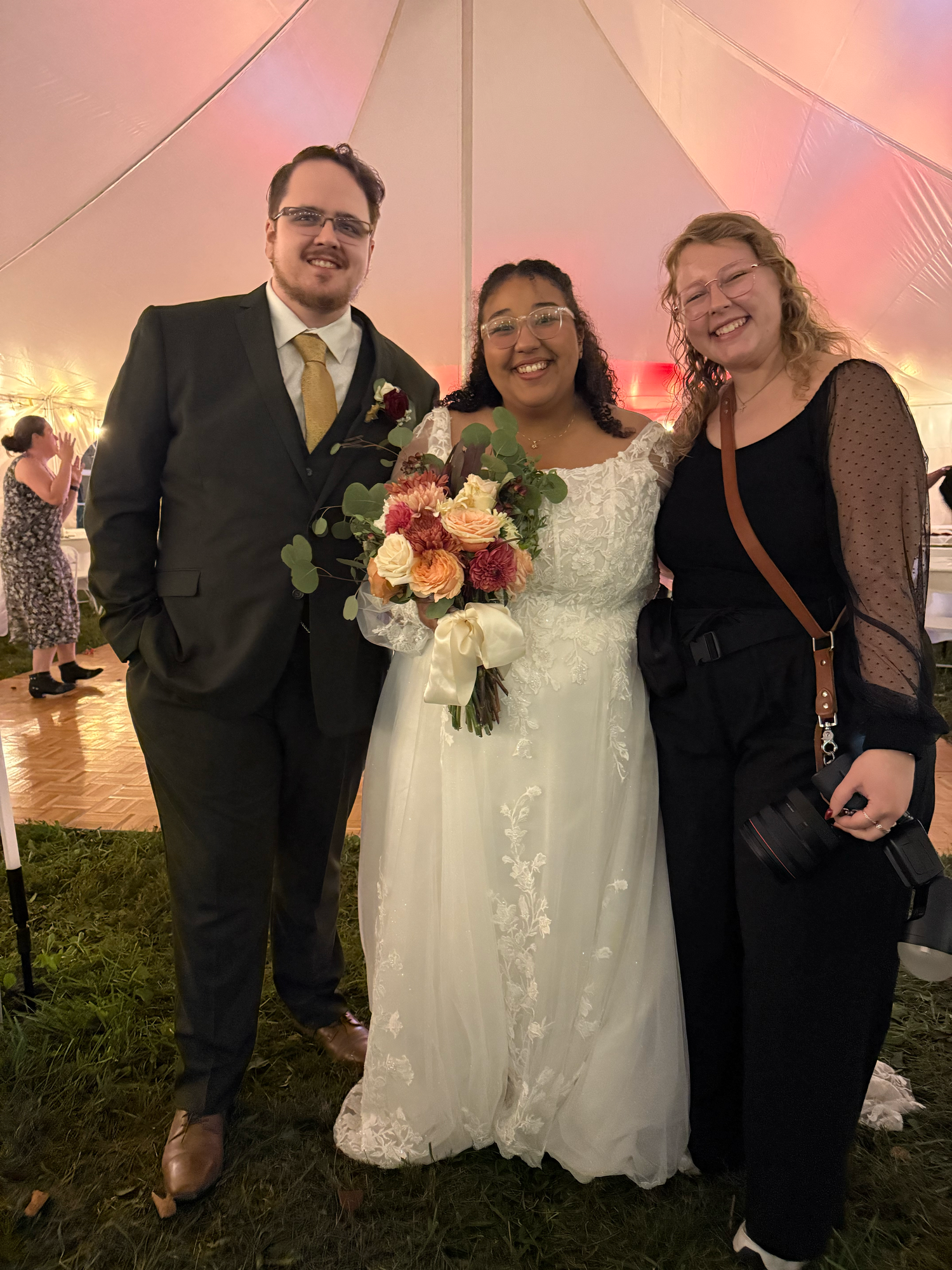 Bride in a white lace gown holding a bouquet poses with a groom in a dark suit and a smiling guest under a tent at an evening wedding.