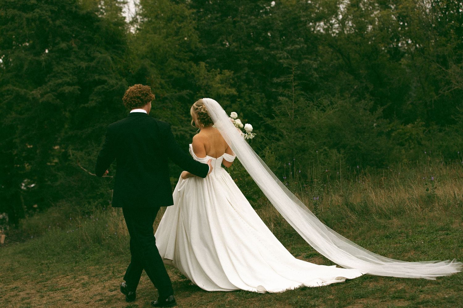 Bride in an off-the-shoulder white wedding gown with a long flowing veil walks hand in hand with a groom in a dark suit through a grassy out