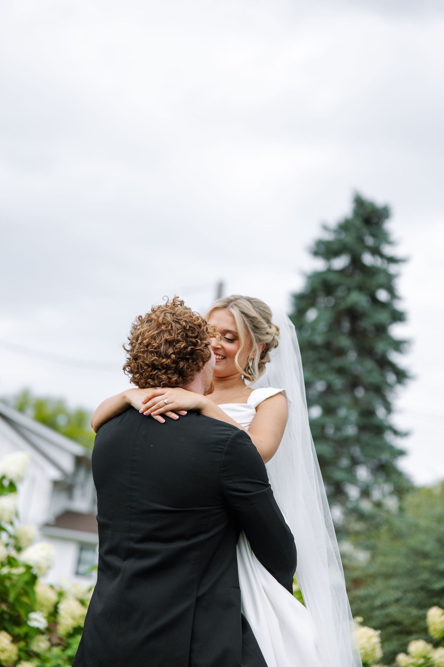 Smiling bride in a white dress and long veil is lifted in an embrace by her curly-haired groom in a black suit outdoors.