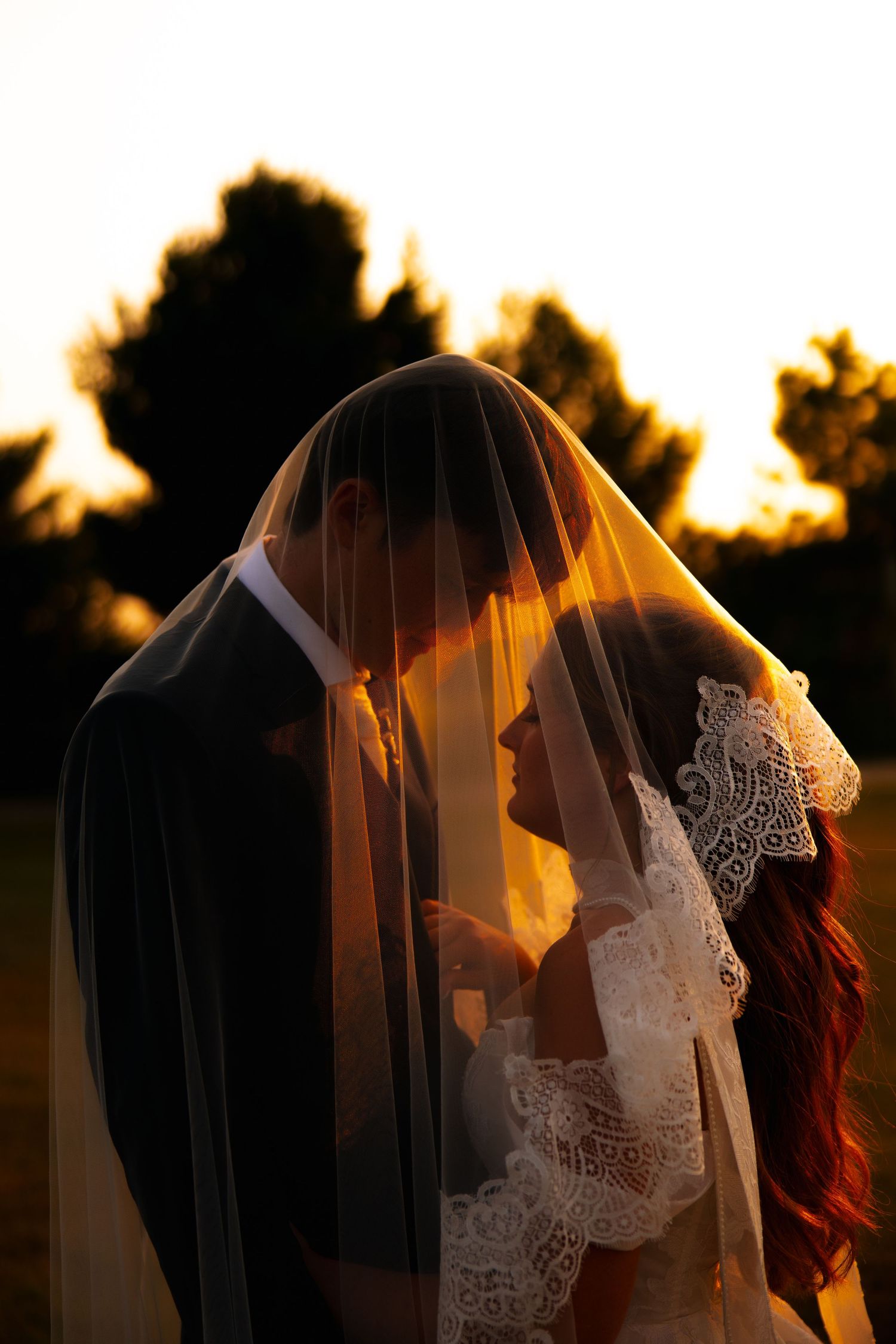Bride and groom share a quiet moment beneath a sheer lace veil glowing in golden sunset light.