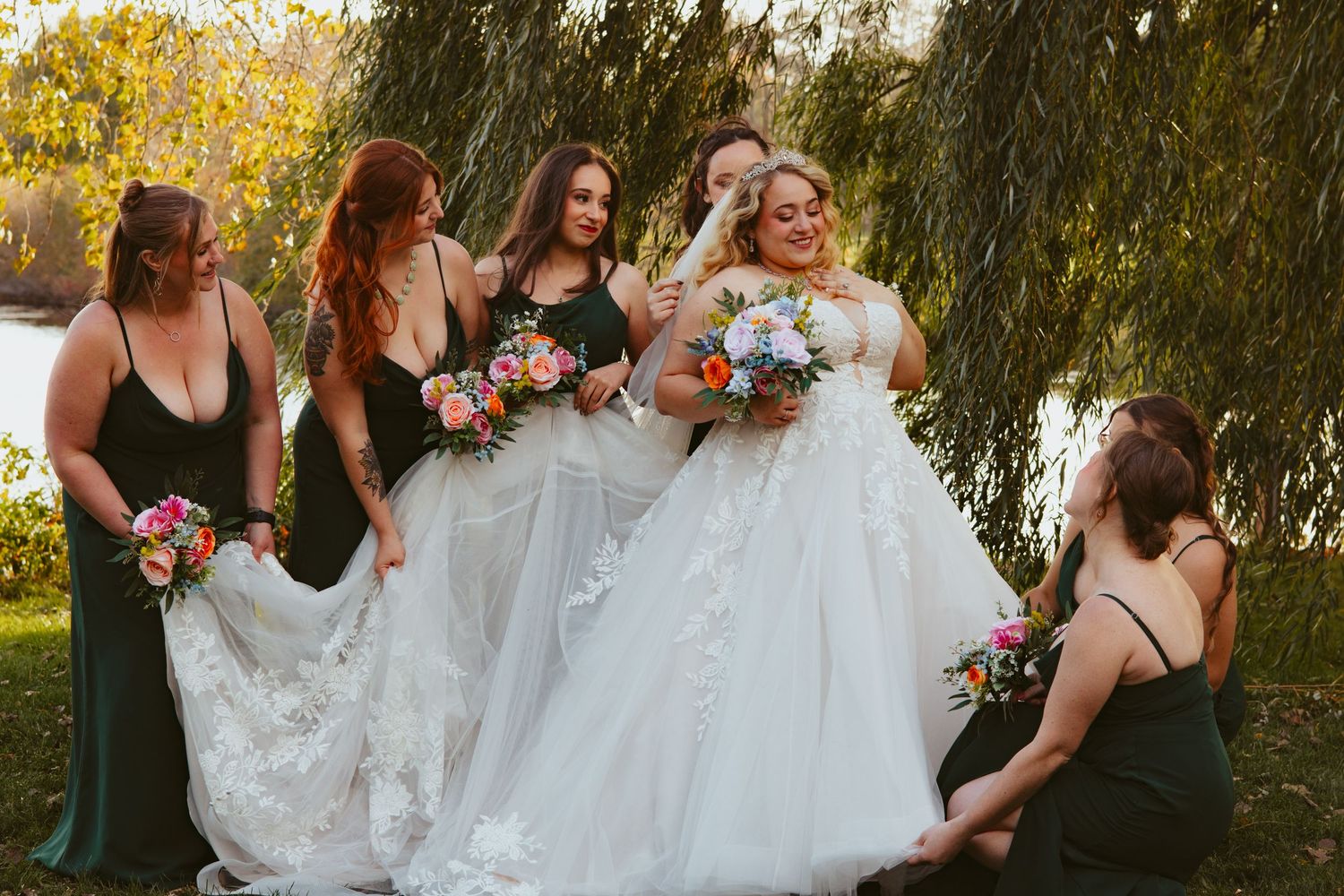Bride in a flowing white lace wedding gown surrounded by bridesmaids in dark green dresses holding bouquets beside a willow tree.