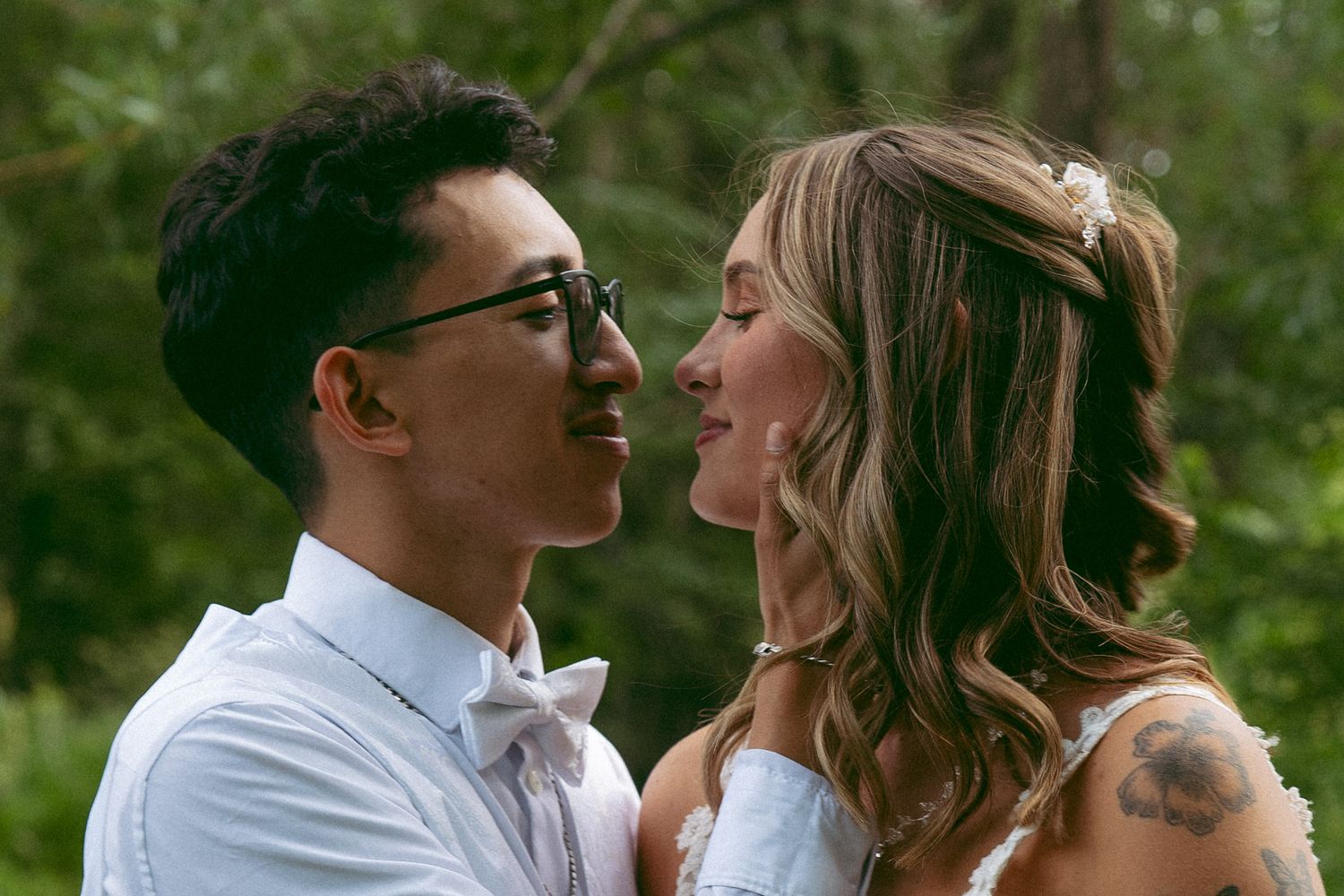 A groom in glasses and a bow tie leans in to kiss a bride with wavy hair and a floral hairpin among green trees.