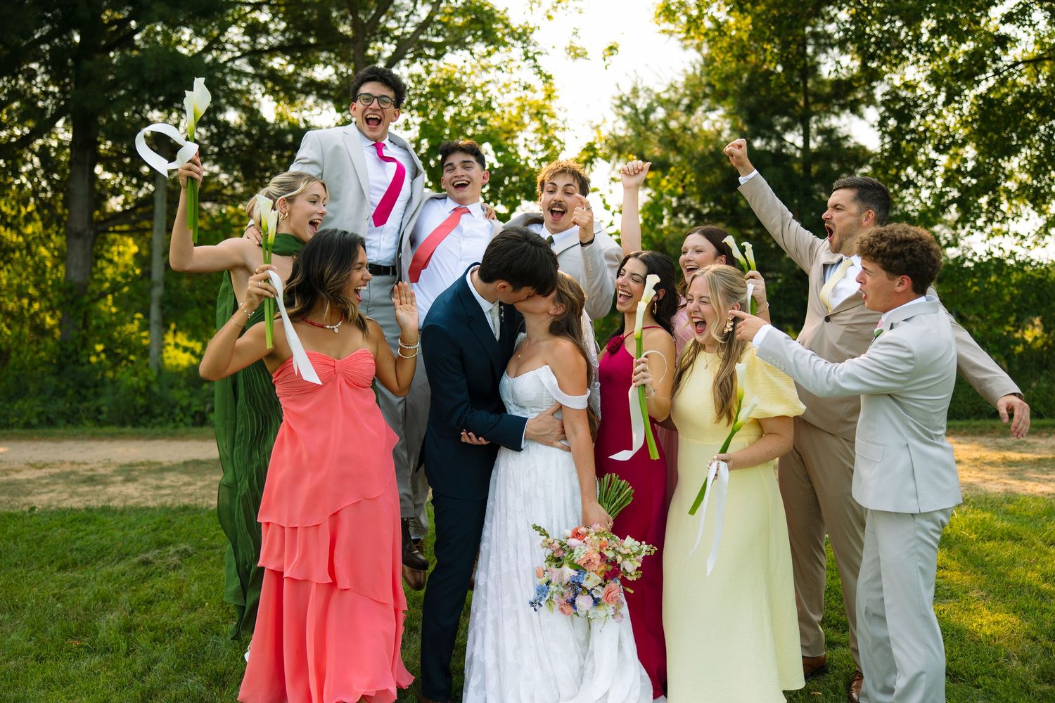 A joyful bridal party cheers and celebrates around a kissing bride and groom on a sunny lawn, with one guest holding up a giant key.