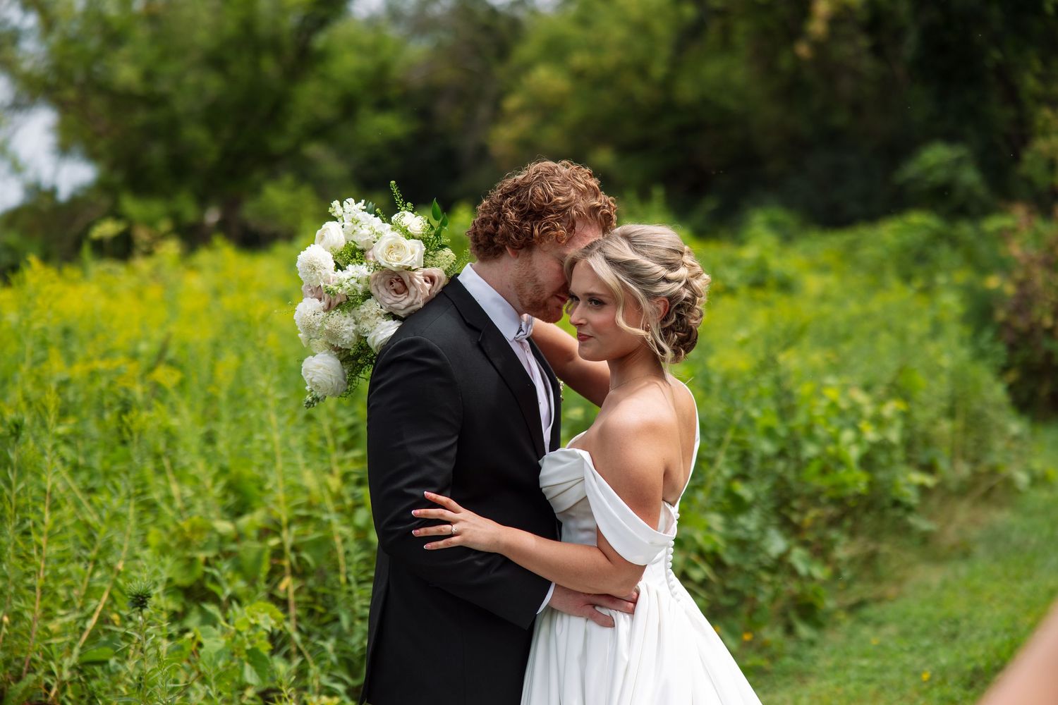 Bride in an off-shoulder white gown and groom in a dark suit embrace foreheads together holding a white bouquet in a green summer field.