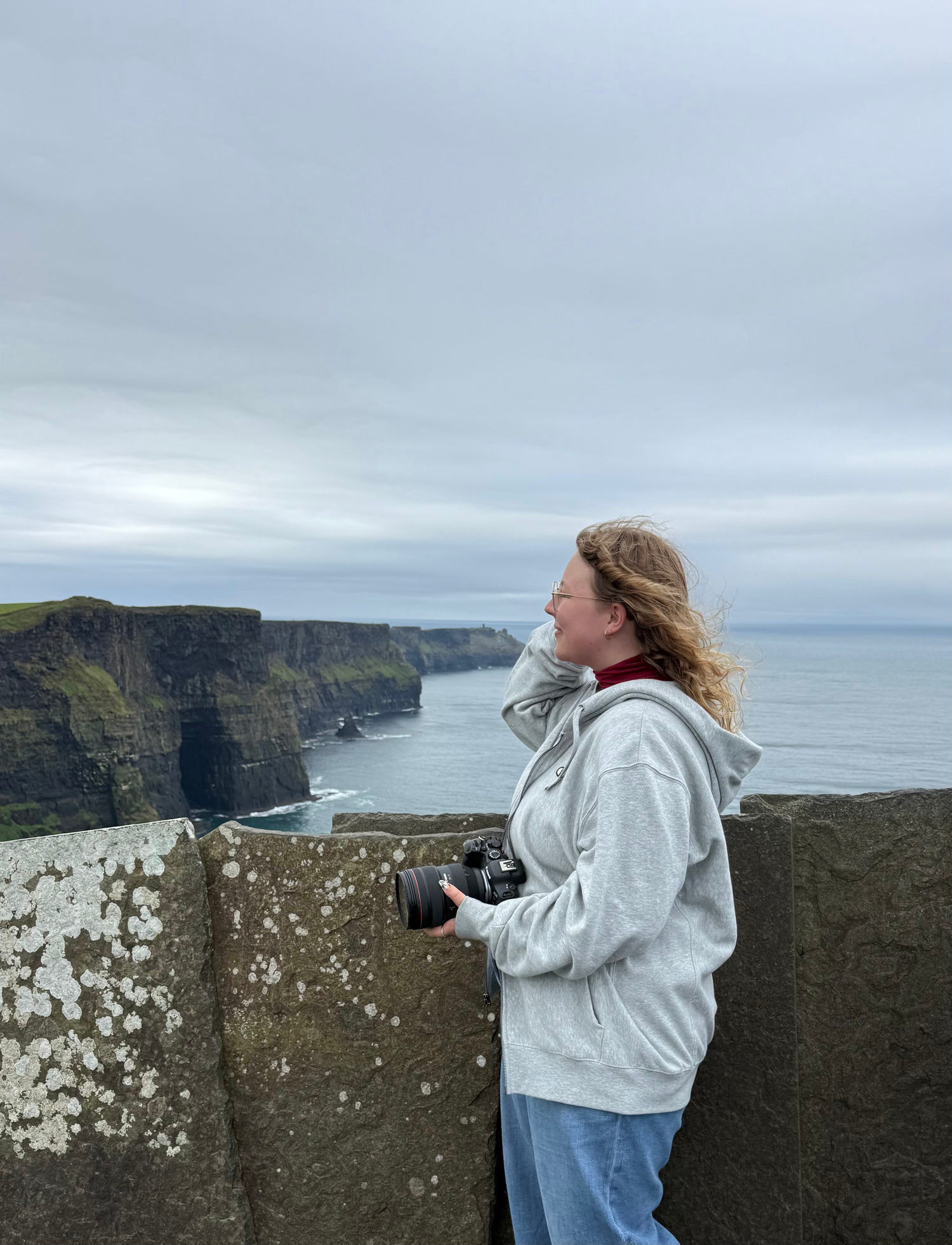 Woman in a gray hoodie holding a camera and gazing out from a stone wall toward dramatic sea cliffs under an overcast sky.