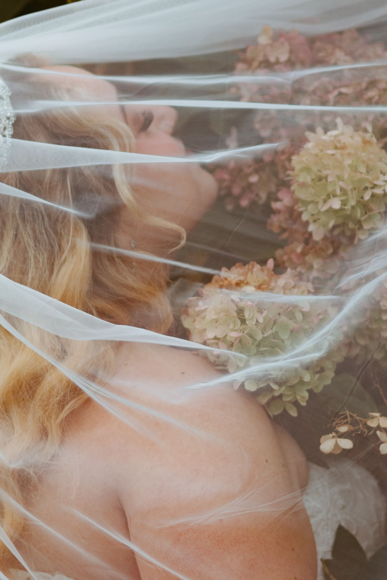 Bride with wavy blonde hair and bare shoulders rests beneath a sheer veil beside dried pink and green hydrangea blooms.
