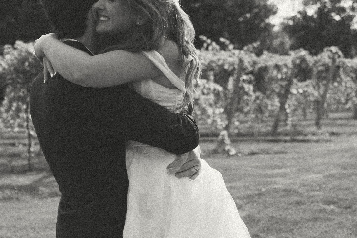 A black and white photo of a bride in a white dress hugging her groom tightly in front of rows of grapevines at a vineyard.