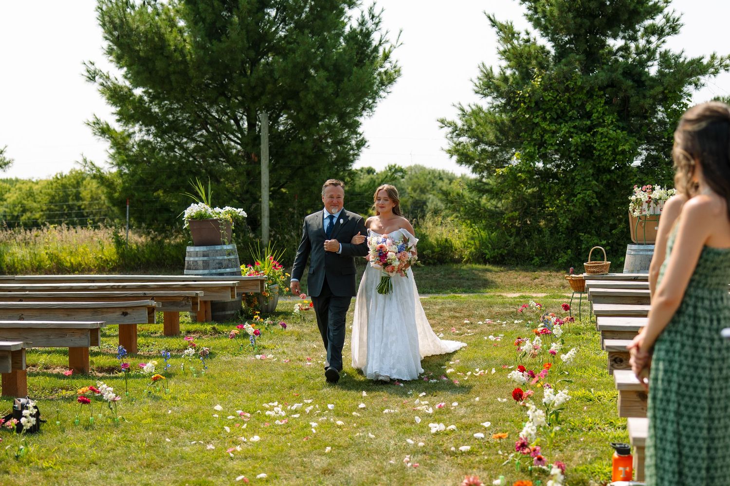 A father walks his bride in a white gown down a grass aisle lined with wooden benches and scattered flower petals at an outdoor wedding.