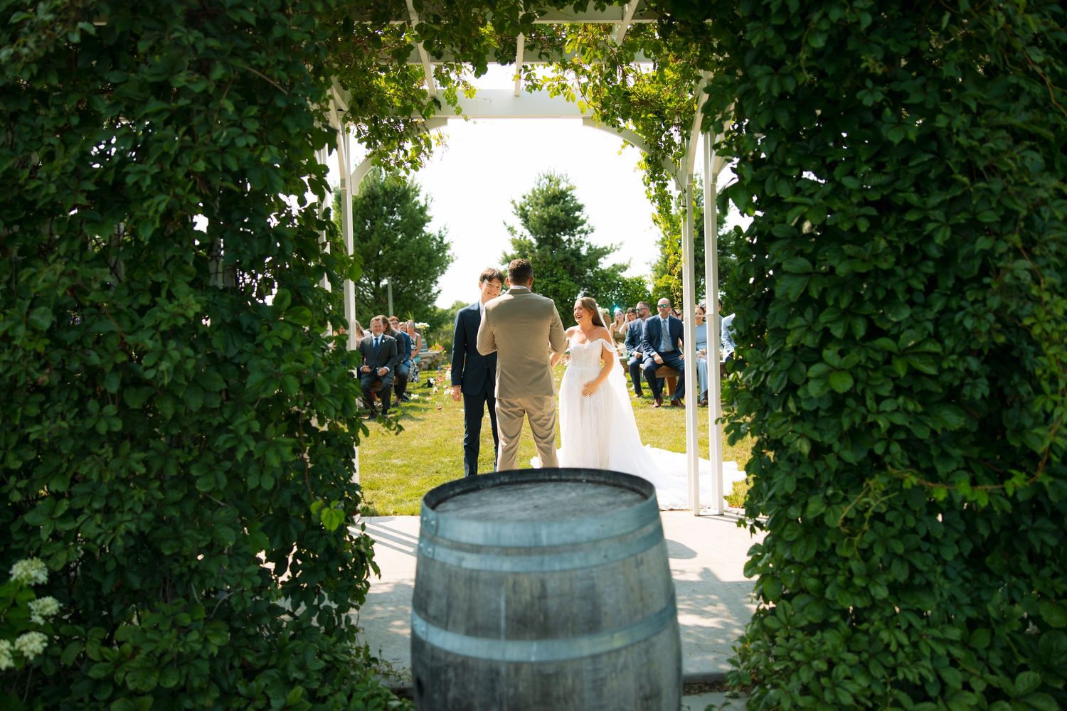 Through a leafy green arbor, a couple stands before wedding guests on the lawn behind a wooden barrel at an outdoor ceremony.