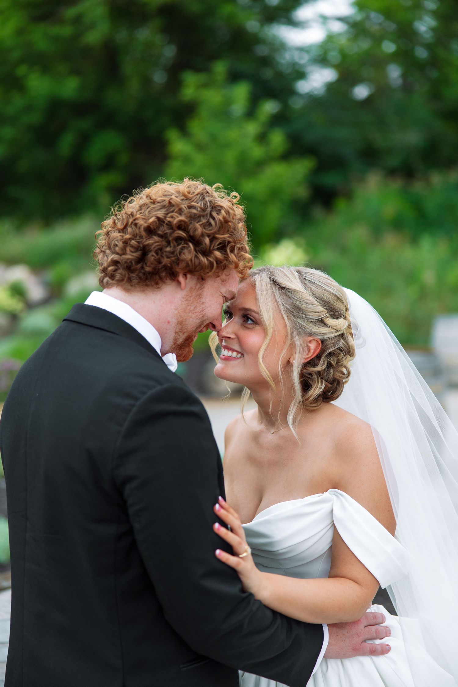 Smiling bride in an off-shoulder white gown and veil embraces her curly red-haired groom in a black tuxedo outdoors.