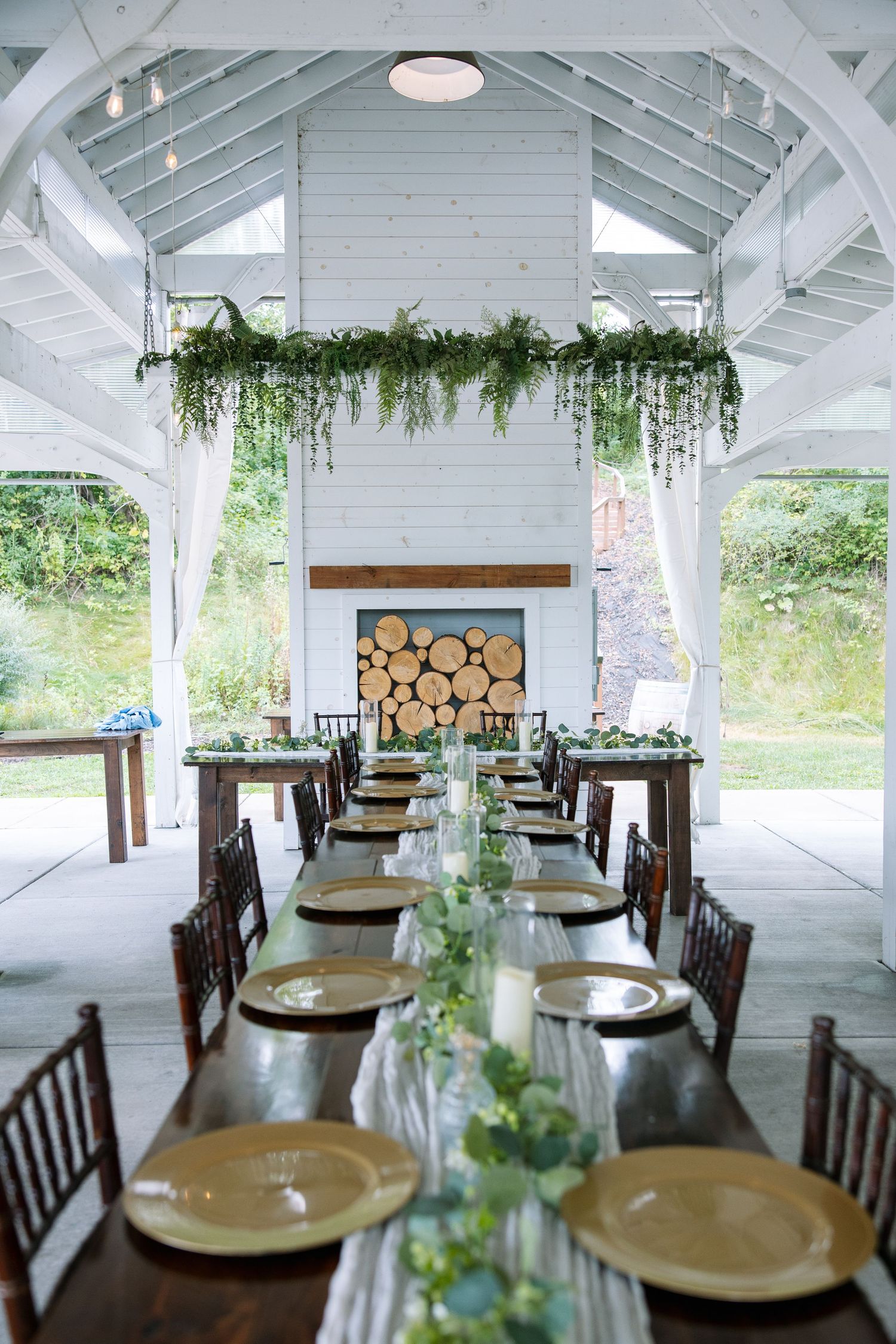 Rustic wedding reception pavilion with long wooden tables, gold charger plates, greenery garland, and a stacked firewood fireplace.