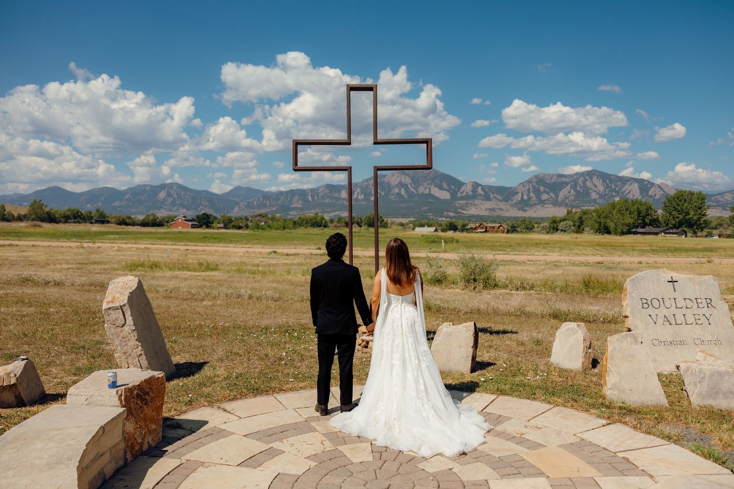 Bride in white gown and groom in dark suit hold hands before a large cross at a stone outdoor chapel with mountain views.