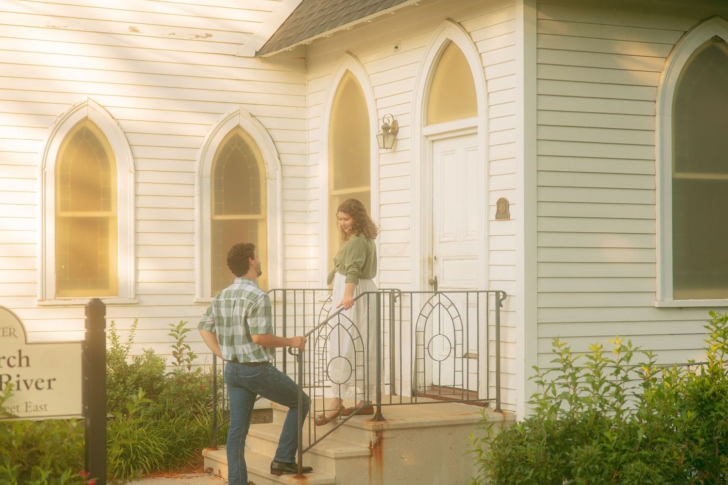 Couple stands at the steps of a charming white wooden chapel in warm golden evening light.