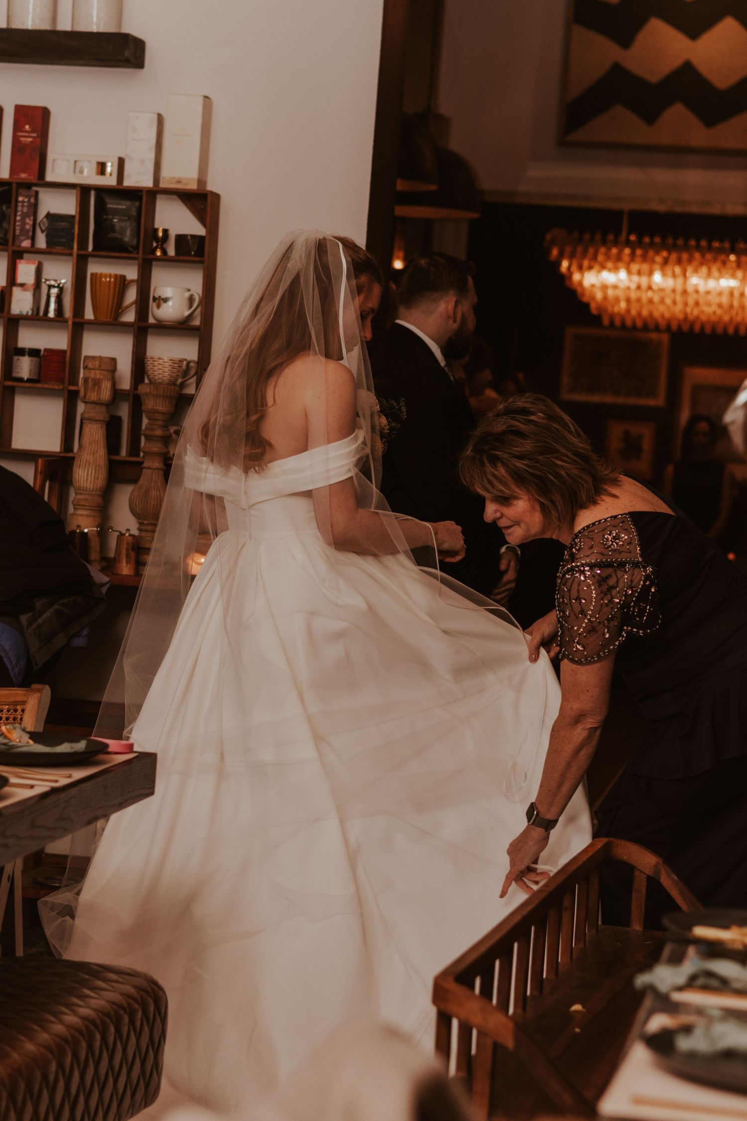 Bride in an off-the-shoulder gown with a long veil as a woman in a beaded dress adjusts her skirt at the reception.