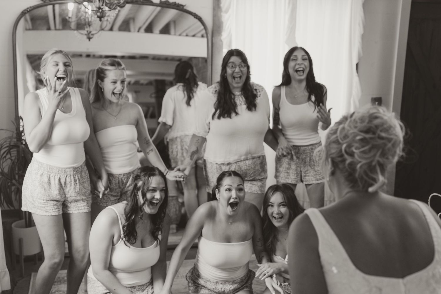 Black and white photo of bridesmaids in matching tops and shorts laughing and cheering as they react to the bride in a bright room.