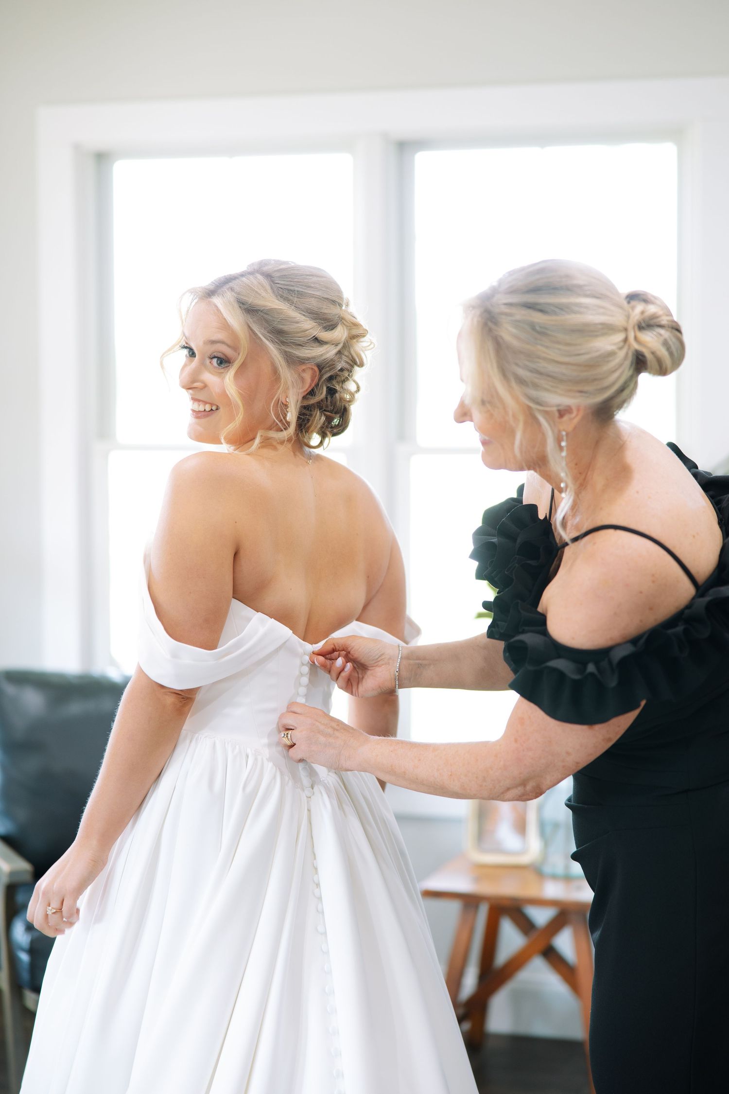 Smiling bride in an off-the-shoulder white wedding gown glances back as a woman in black fastens the buttons.