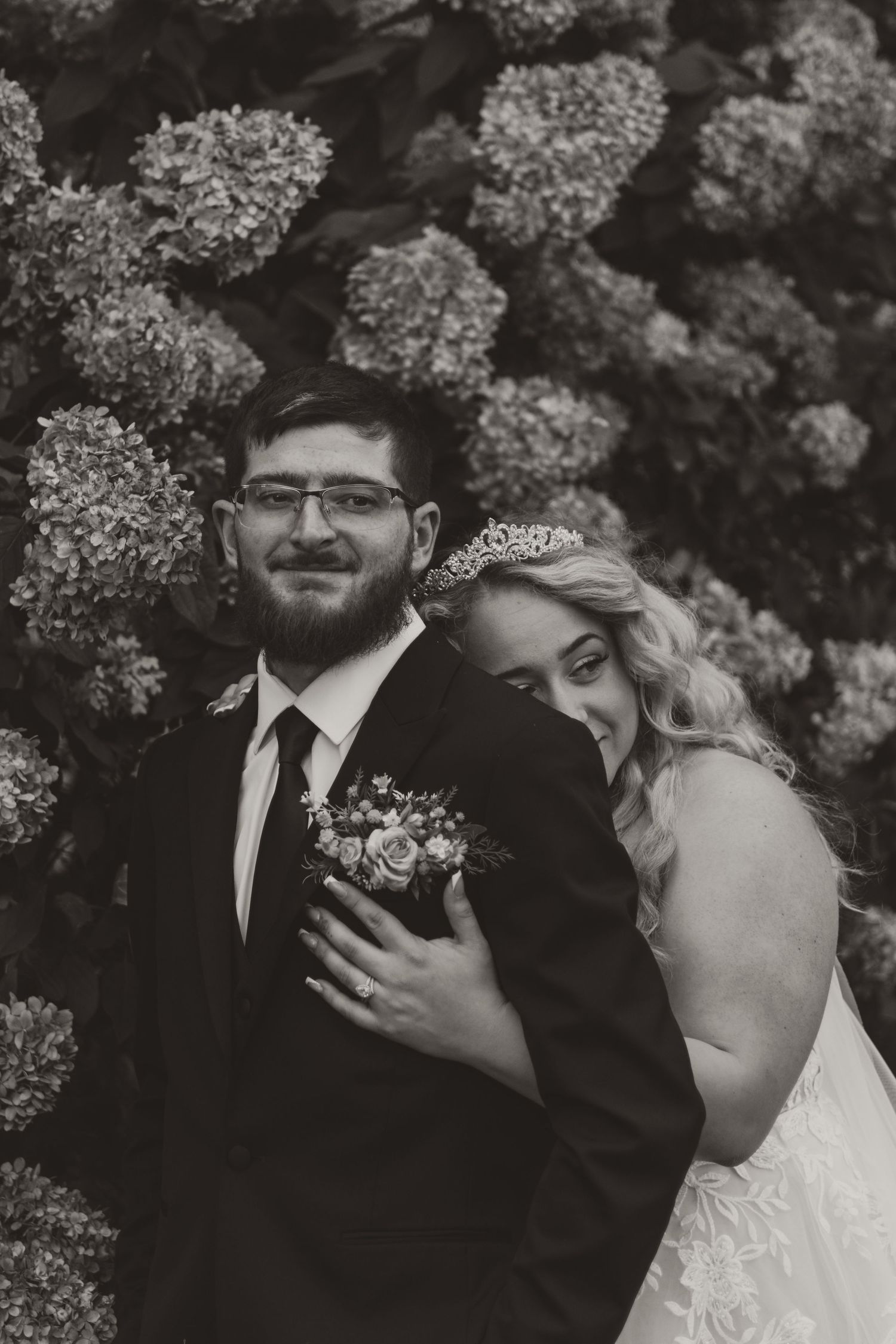 Black and white wedding portrait of a smiling groom in glasses and a suit as his bride in a tiara embraces him before hydrangeas.