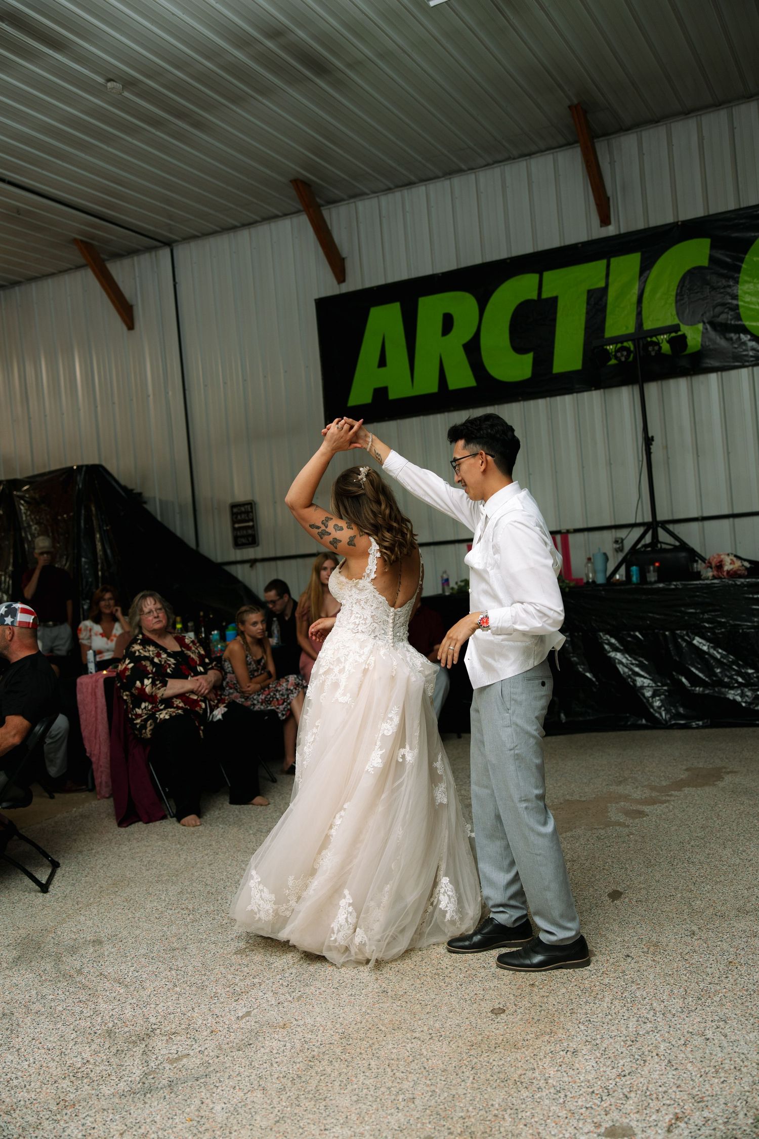 A bride in a lace gown and a groom in gray pants share a first dance at a wedding reception as guests watch.