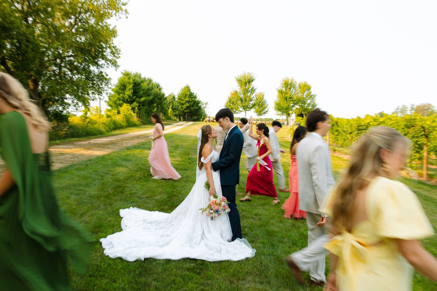 A bride and groom embrace on a grassy path as bridesmaids and groomsmen in pink, green, and yellow walk past at a vineyard wedding.