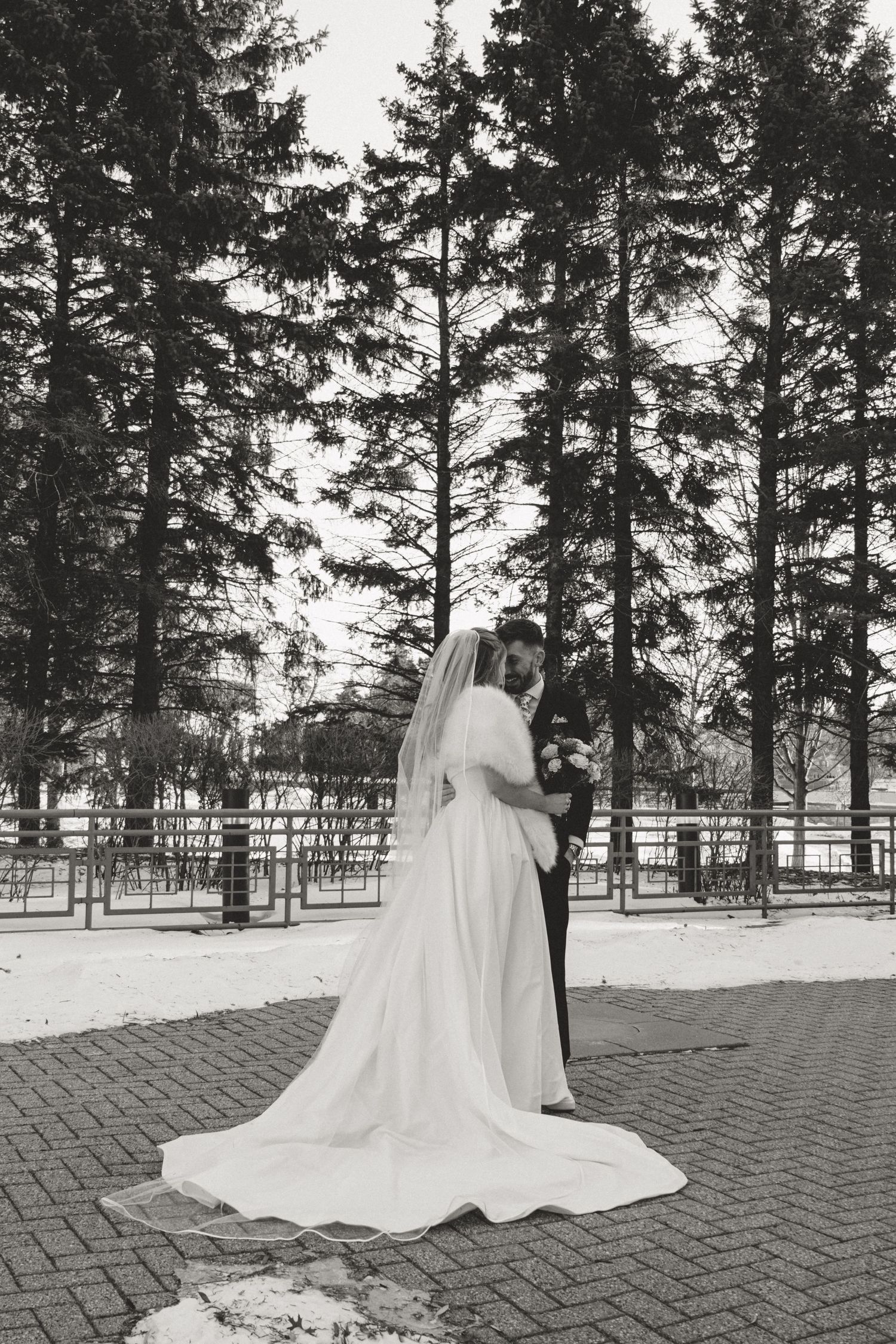 Black and white winter wedding portrait of a bride and groom embracing among tall evergreens with a long gown train.