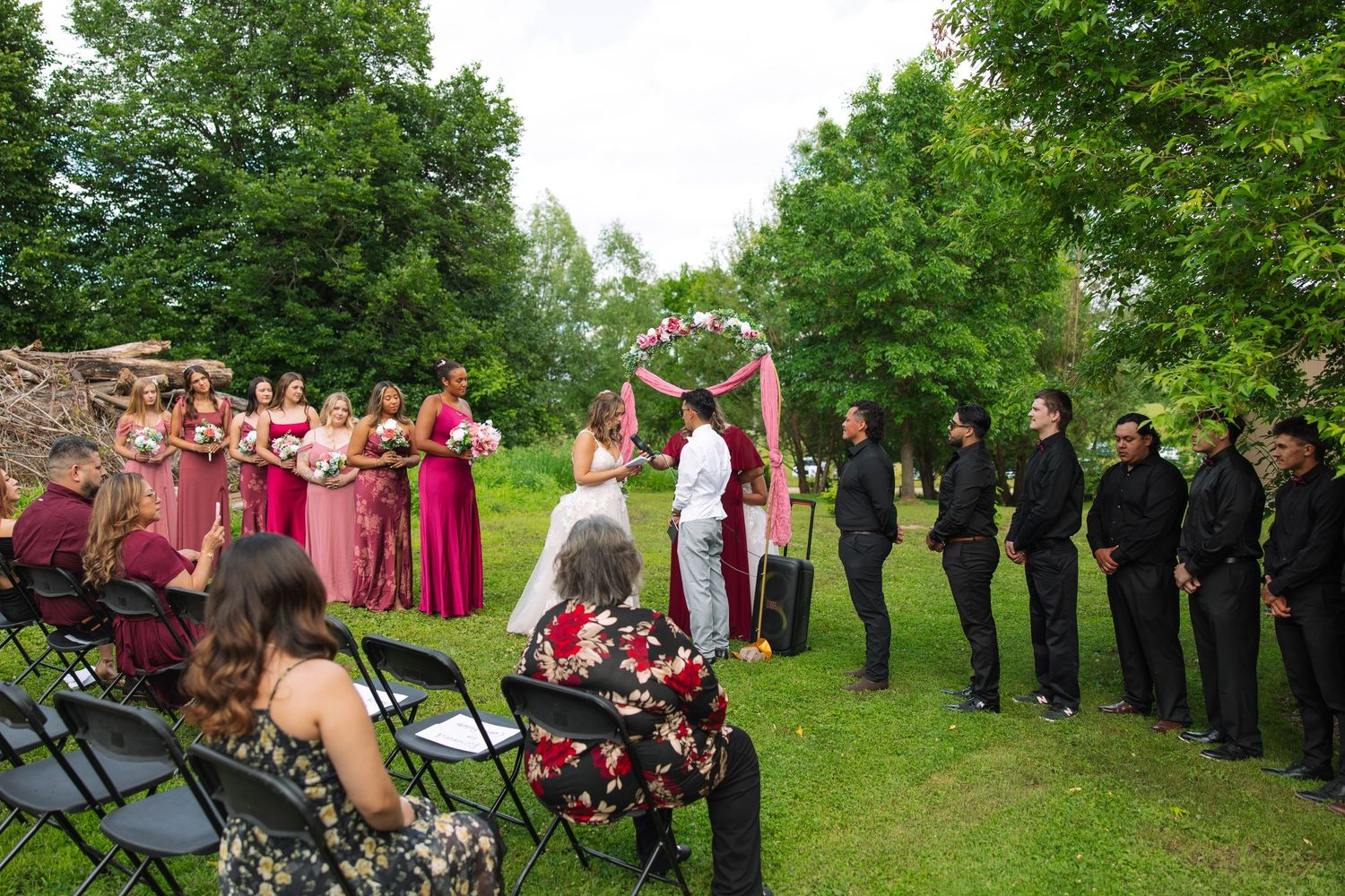 Wedding party and seated guests watch an outdoor ceremony under a floral arch with pink draping on a green lawn among trees.