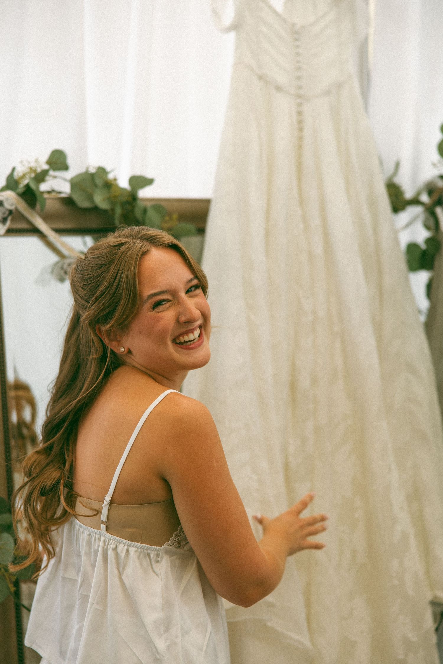 Smiling bride in a white camisole and robe laughs while standing beside her lace wedding dress hanging in a bright room.