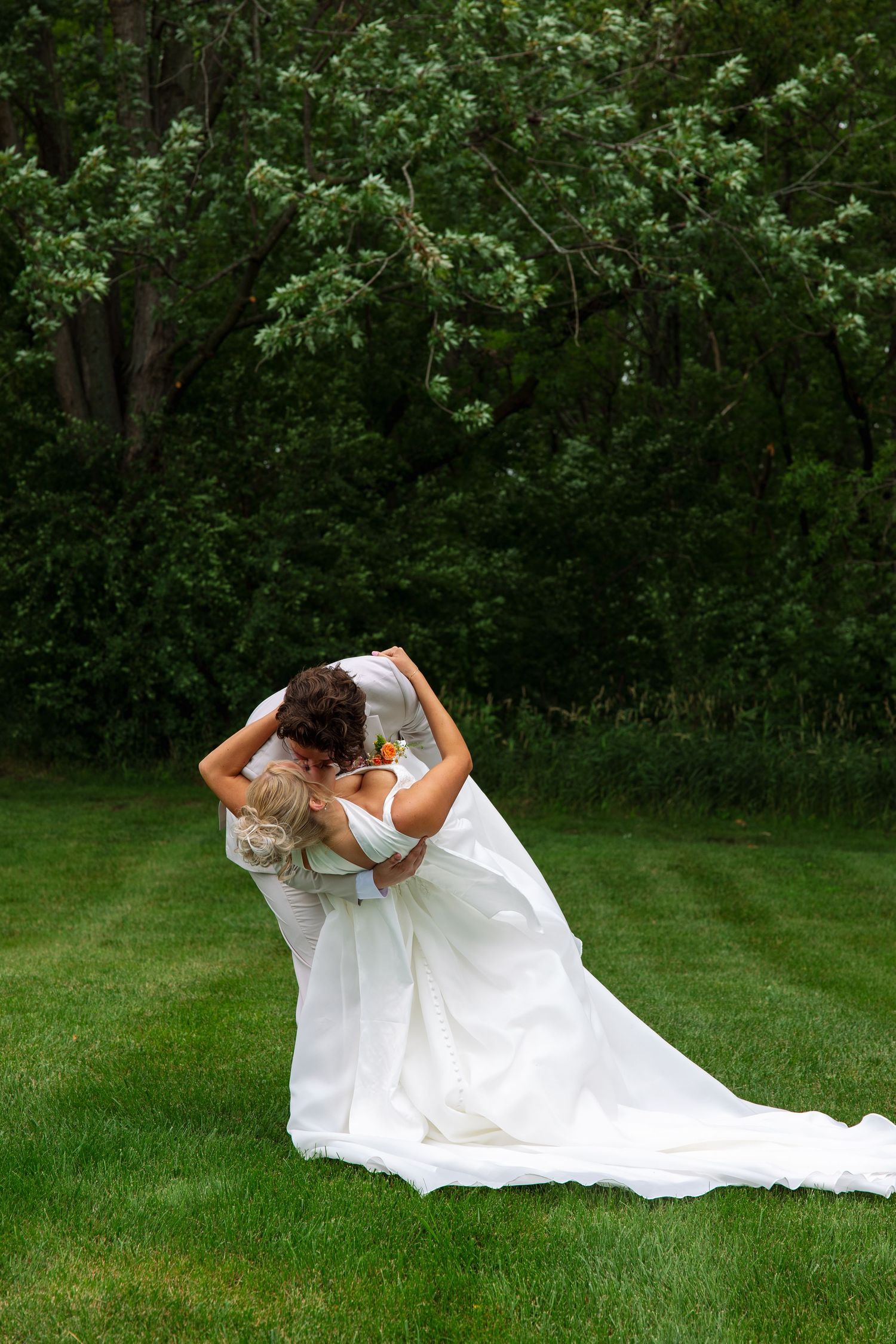 Groom dips the bride in an romantic kiss on a manicured lawn, her long white wedding dress trailing behind them.