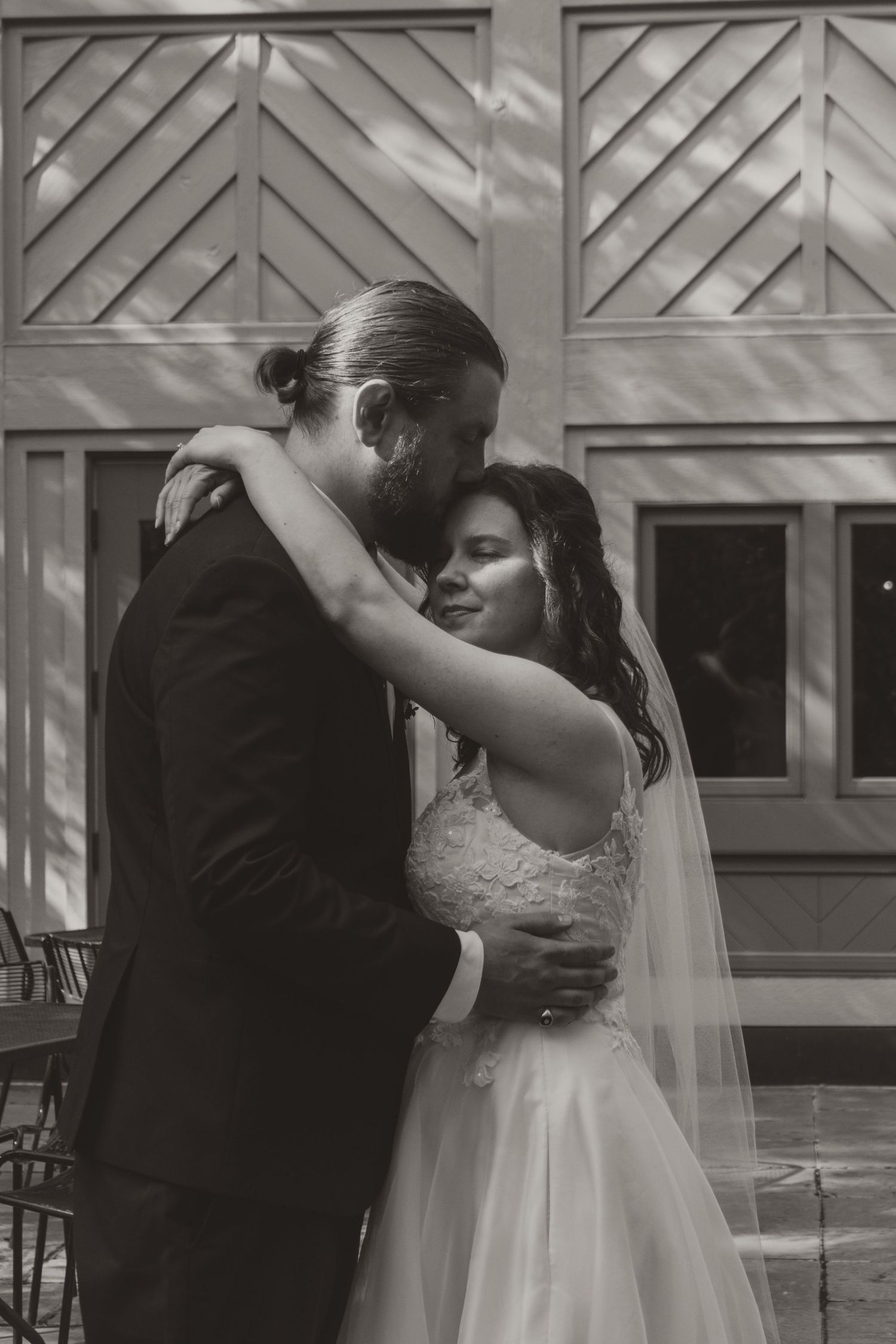 Black and white photo of a bride in a lace gown and veil embraced by a bearded groom in a dark suit during a wedding portrait.