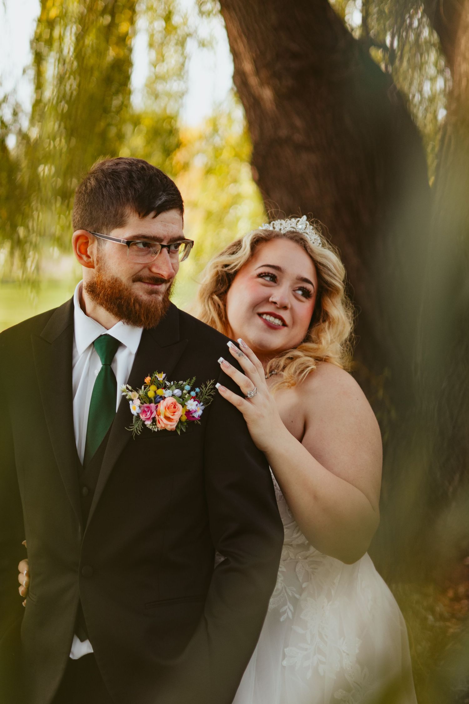 Smiling bride in a lace gown and tiara leans against her bearded groom, who wears glasses, a dark suit, green tie and floral boutonniere.